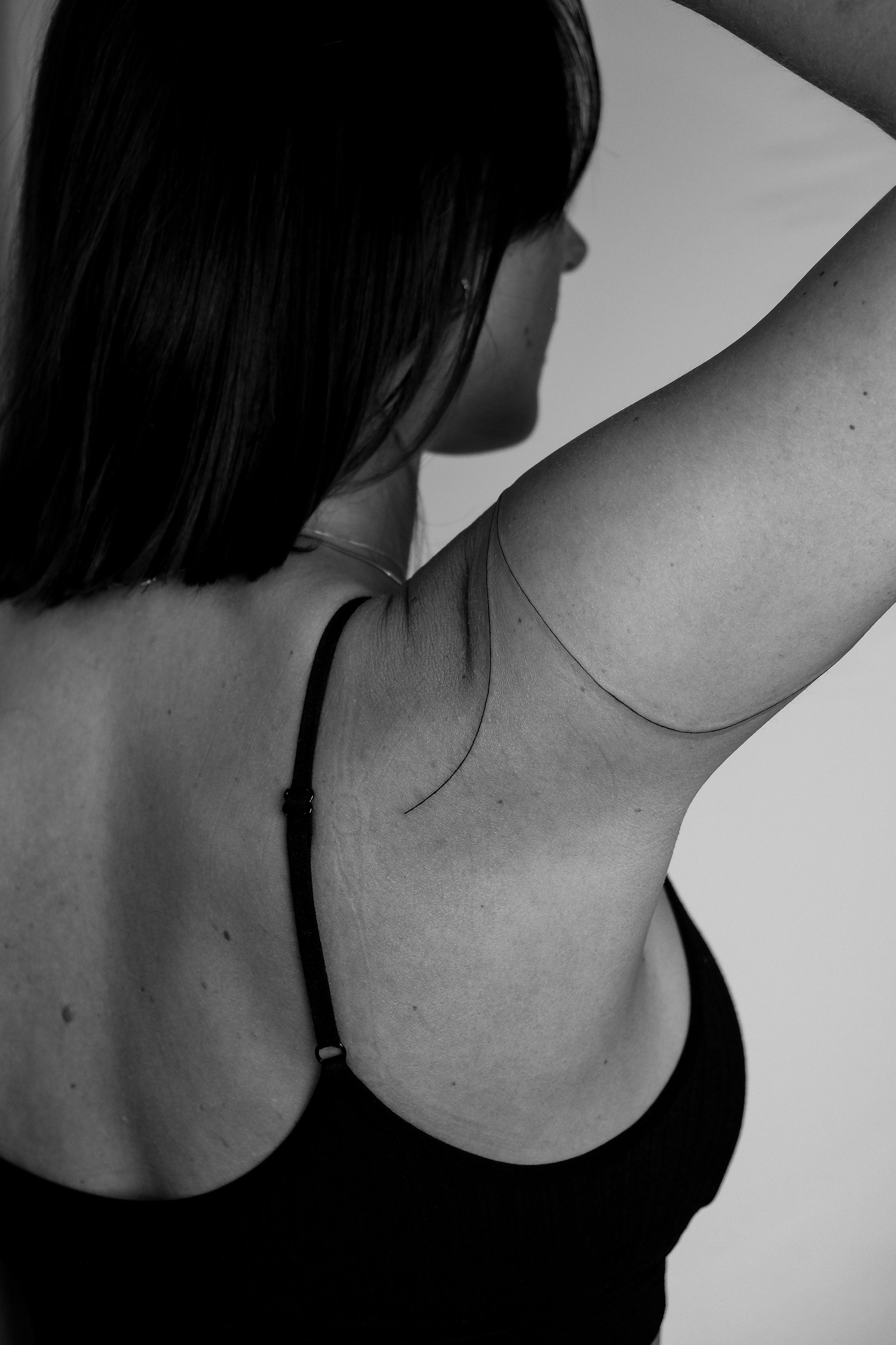 Schwarze und weiße Nahaufnahme einer Frau mit Arm über Kopf, sichtbarer Schulter, Träger eines schwarzen Tops, dunkles Haar, ungeklärter Gesichtsausdruck.