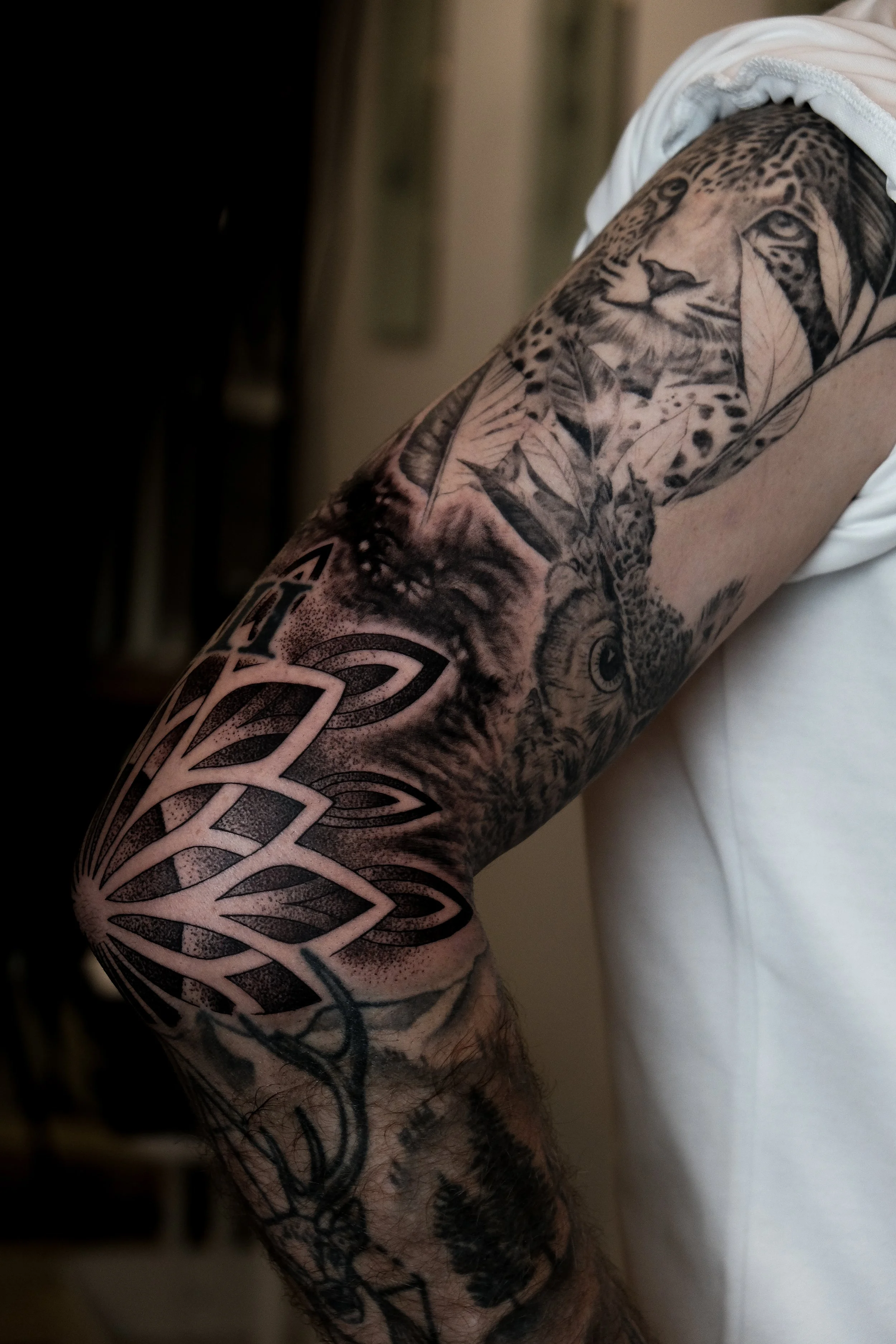 Der Oberarm einer Person mit mehreren schwarzen Tattoos, darunter ein Leopard, eine Eule und geometrische Muster.