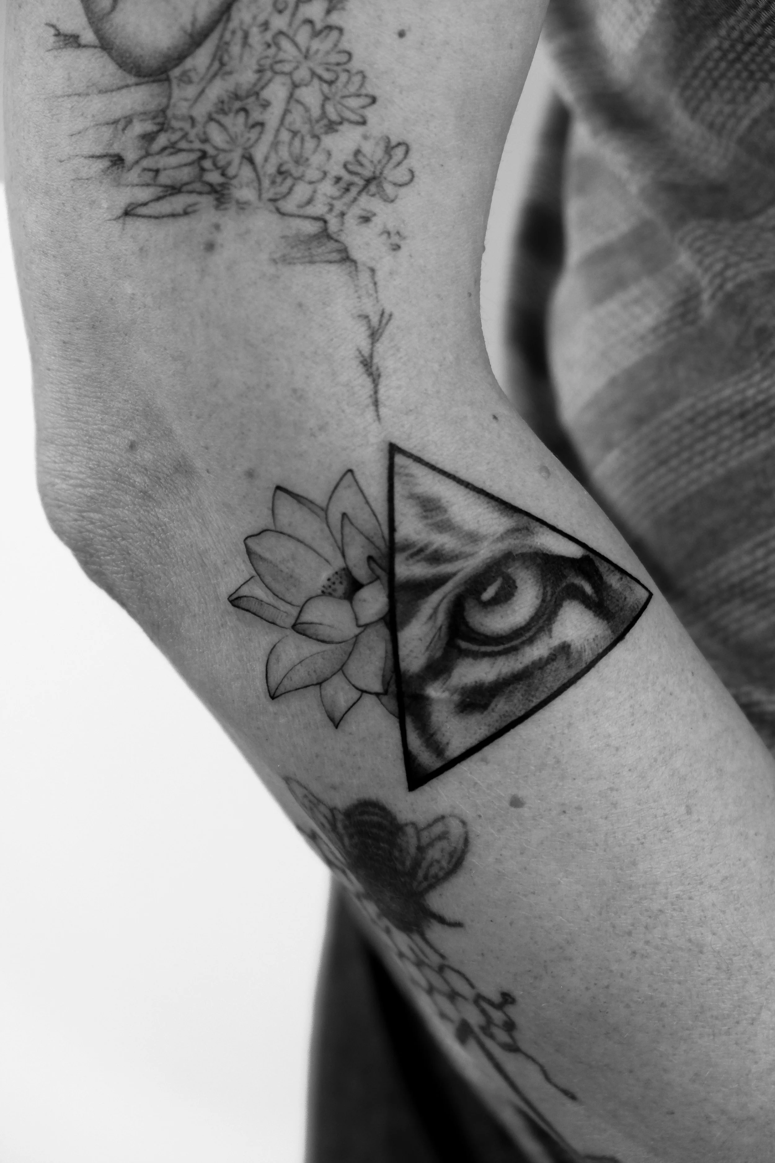 Schwarz-weiße Tätowierungen am Oberarm, inklusive eines Auges innerhalb eines Dreiecks, einer Lotusblume und eines Blümchens.