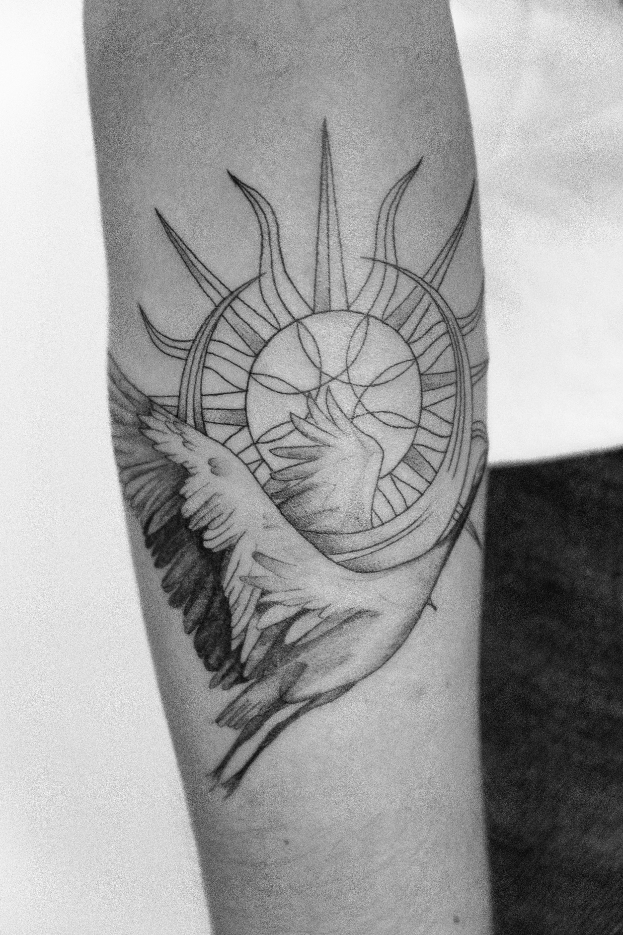 Schwarze Tätowierung auf dem Arm, die eine rosafarbene Sonne mit Strahlen, einen Kreis mit Linien und einen fliegenden Vogel darstellt.