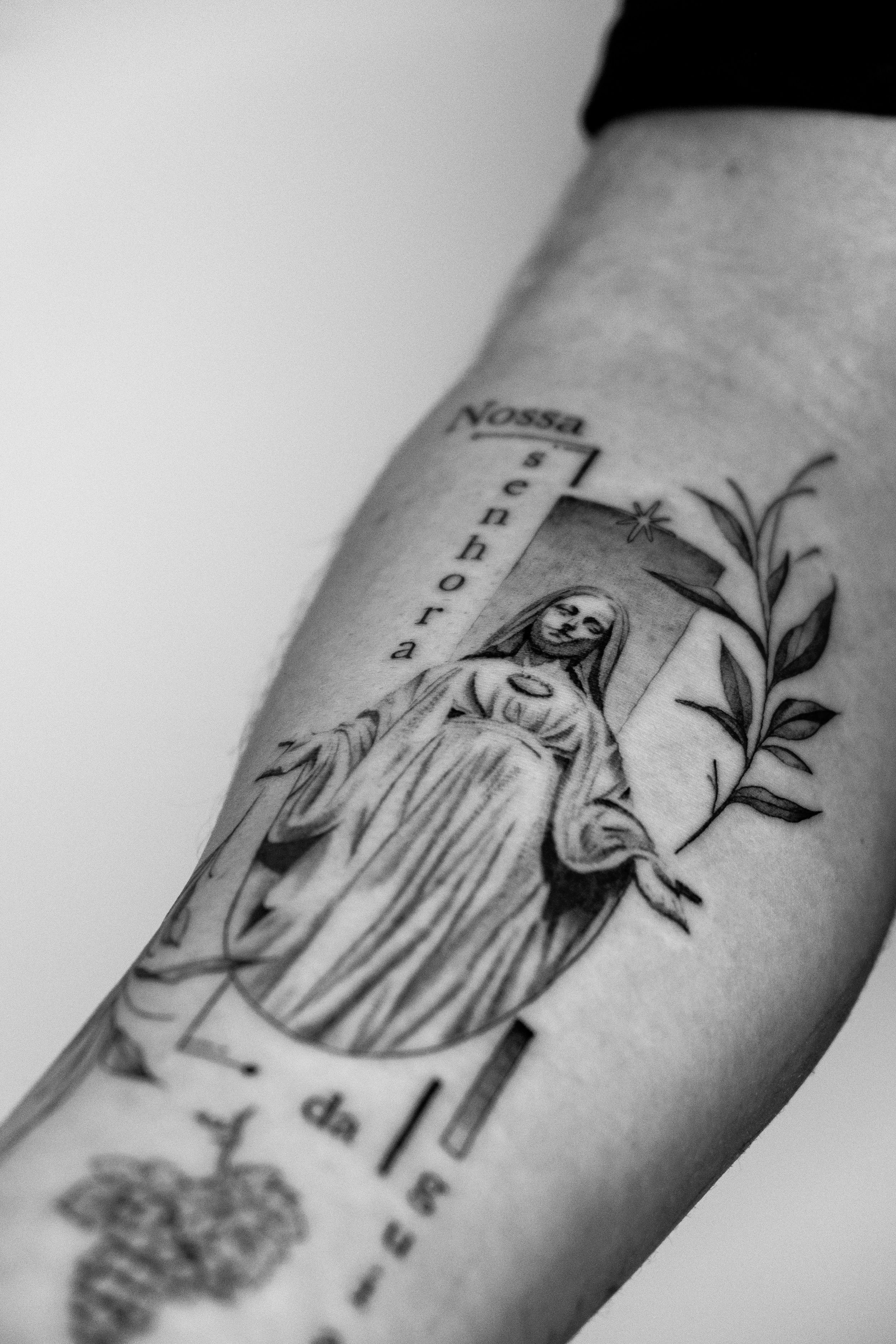 Schwarz-weiß-Tattoo auf einem Arm, zeigt eine Figur mit Heiligenschein, umgeben von Pflanzen und Text in Portugiesisch.