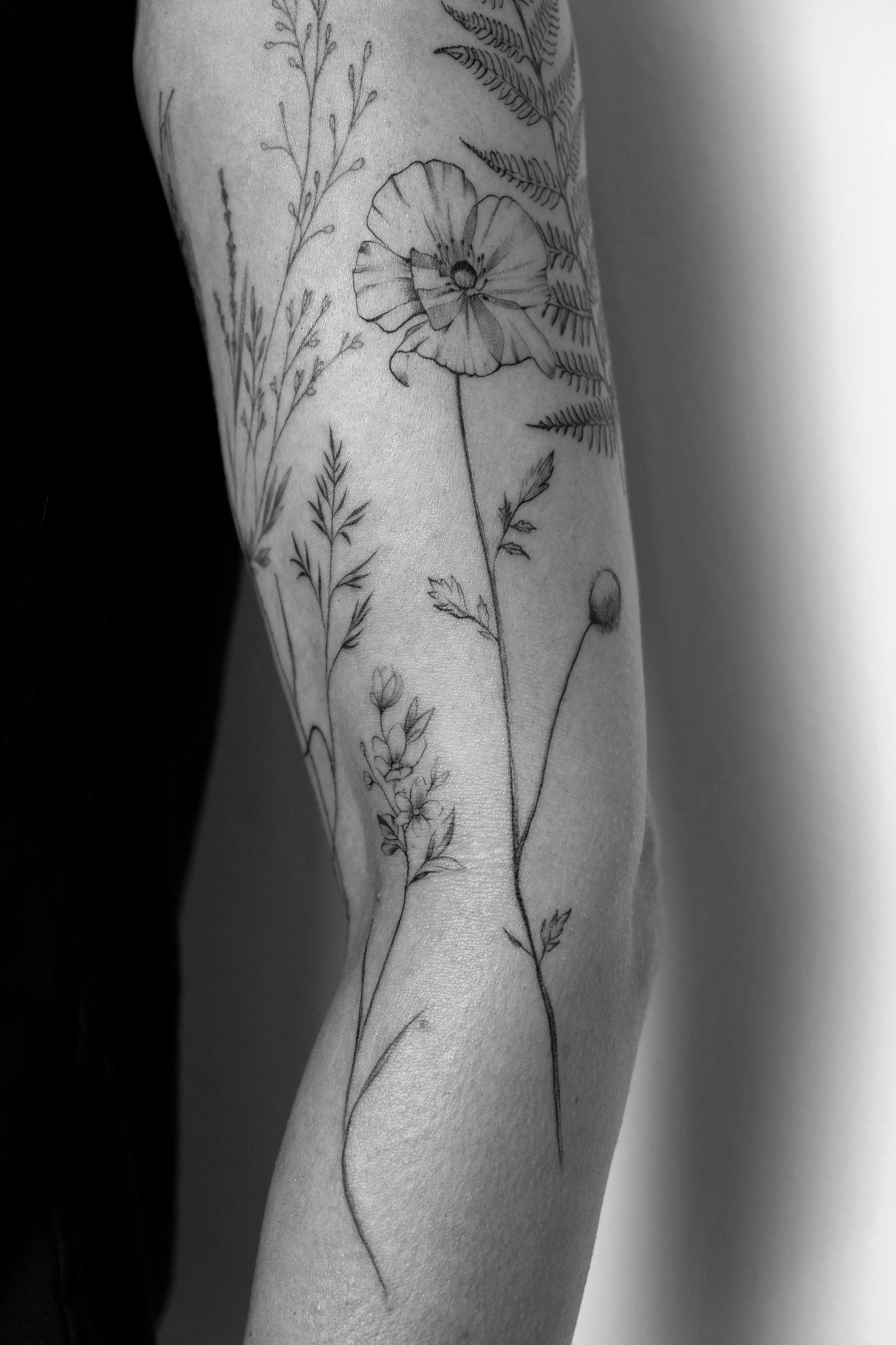 Schwarz-weiß-Tattoo eines Blumenmusters auf einem Arm, das verschiedene Pflanzen und Blumen zeigt.