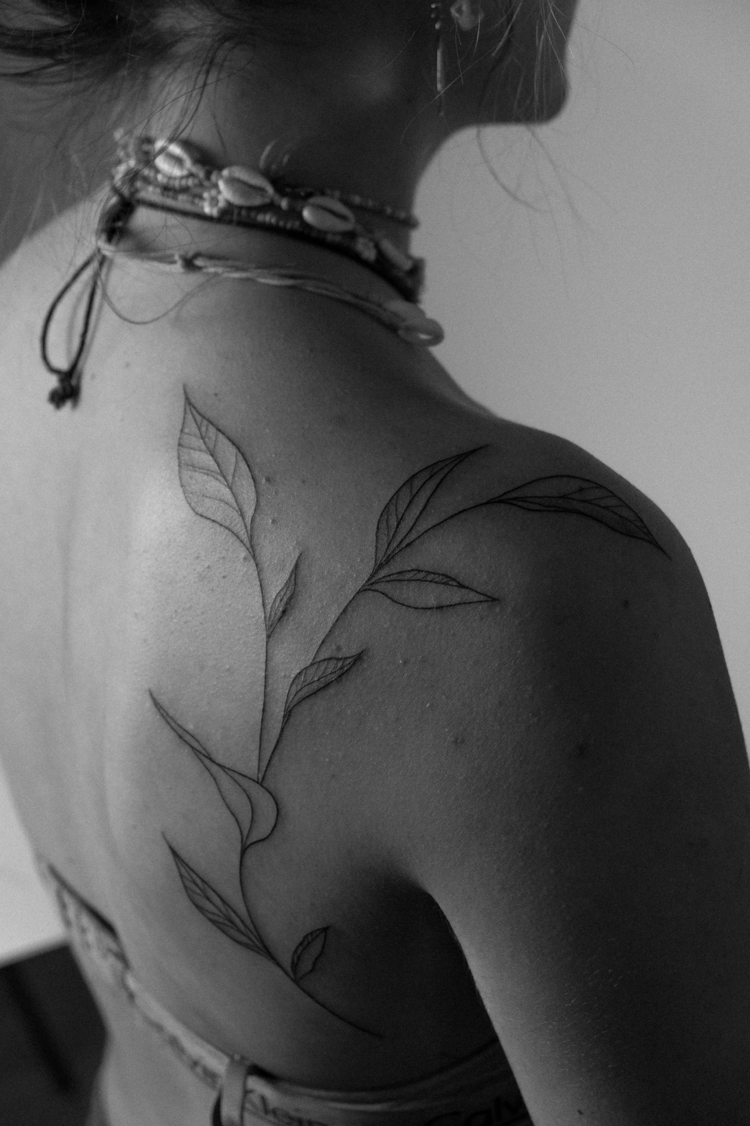 Schulter- und Nacken tattoo einer Person mit Blattmotiv, getragenes Schmuckaccessoire um den Hals, schwarze und weiße Fotografie.