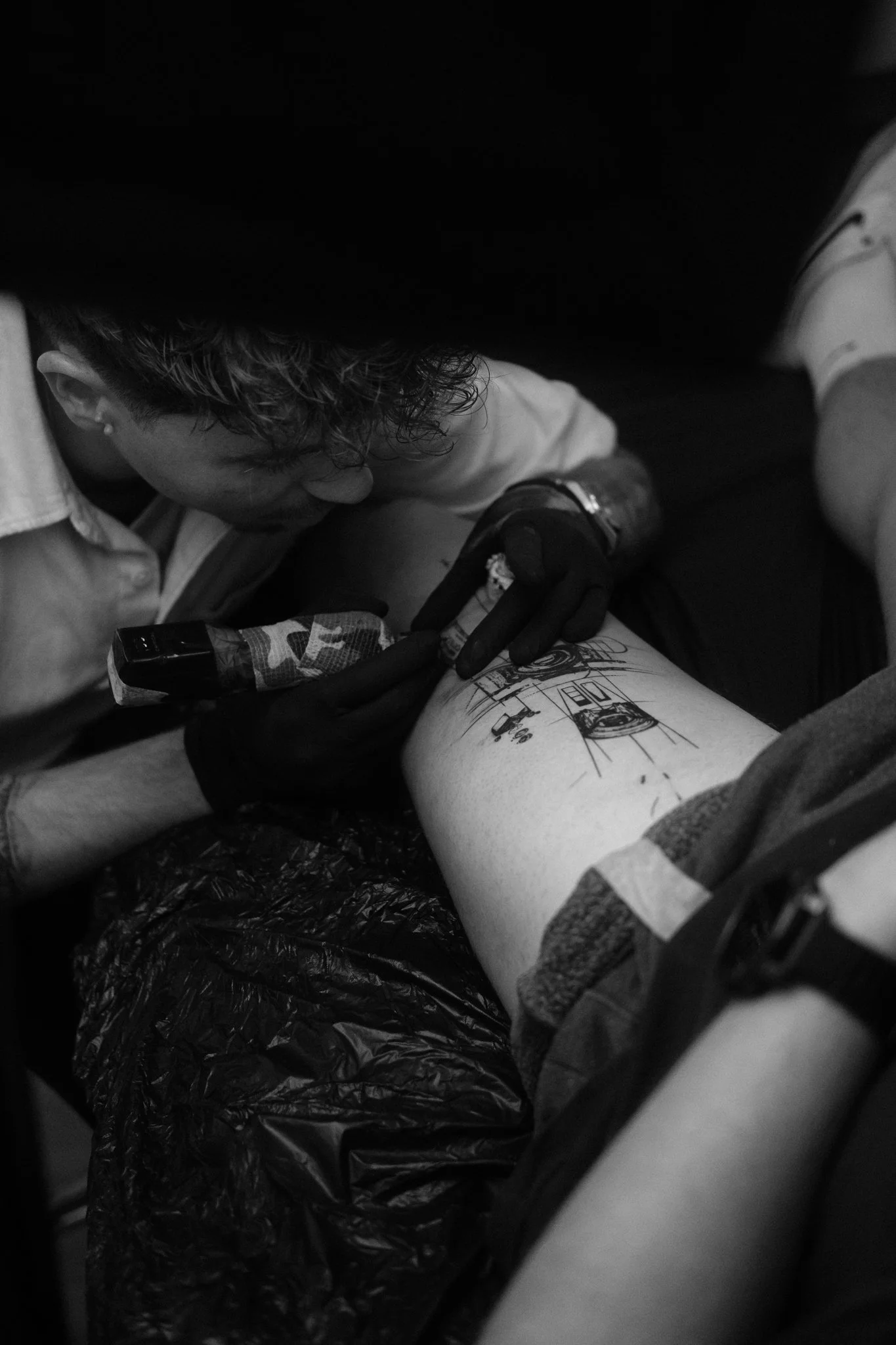 Ein Tätowierer arbeitet an einem Bein eines Kunden im Tattoo Studio Sketchy Riedi in Chur, der auf einer Liege liegt. Die Szene ist in schwarz-weiß.