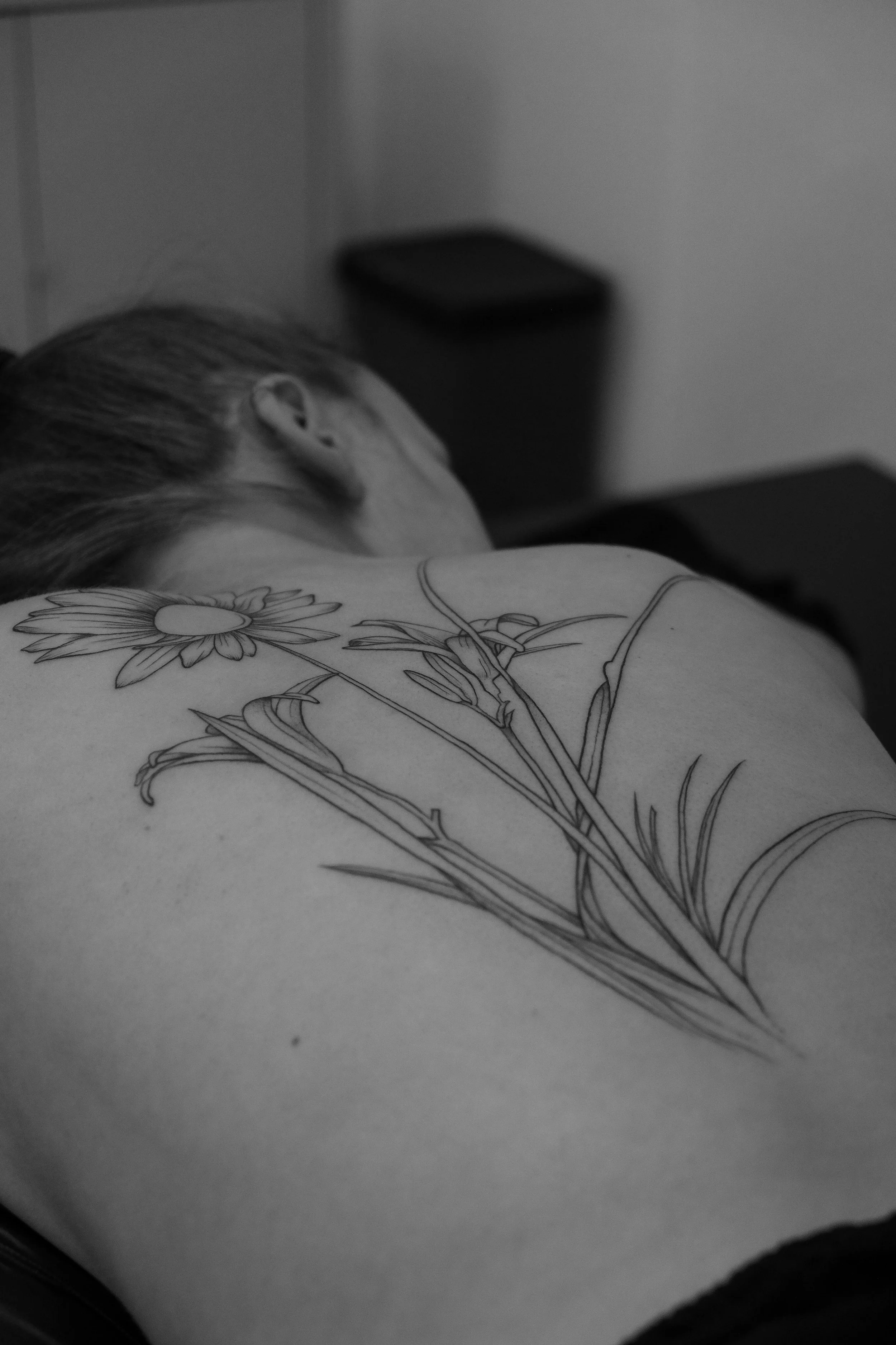 Schwarz-Weiß-Foto einer schlafenden Person mit einer Kerze im Hintergrund, die auf einem Tisch steht, und einem Tattoo eines Blumenmusters auf der Schulter.