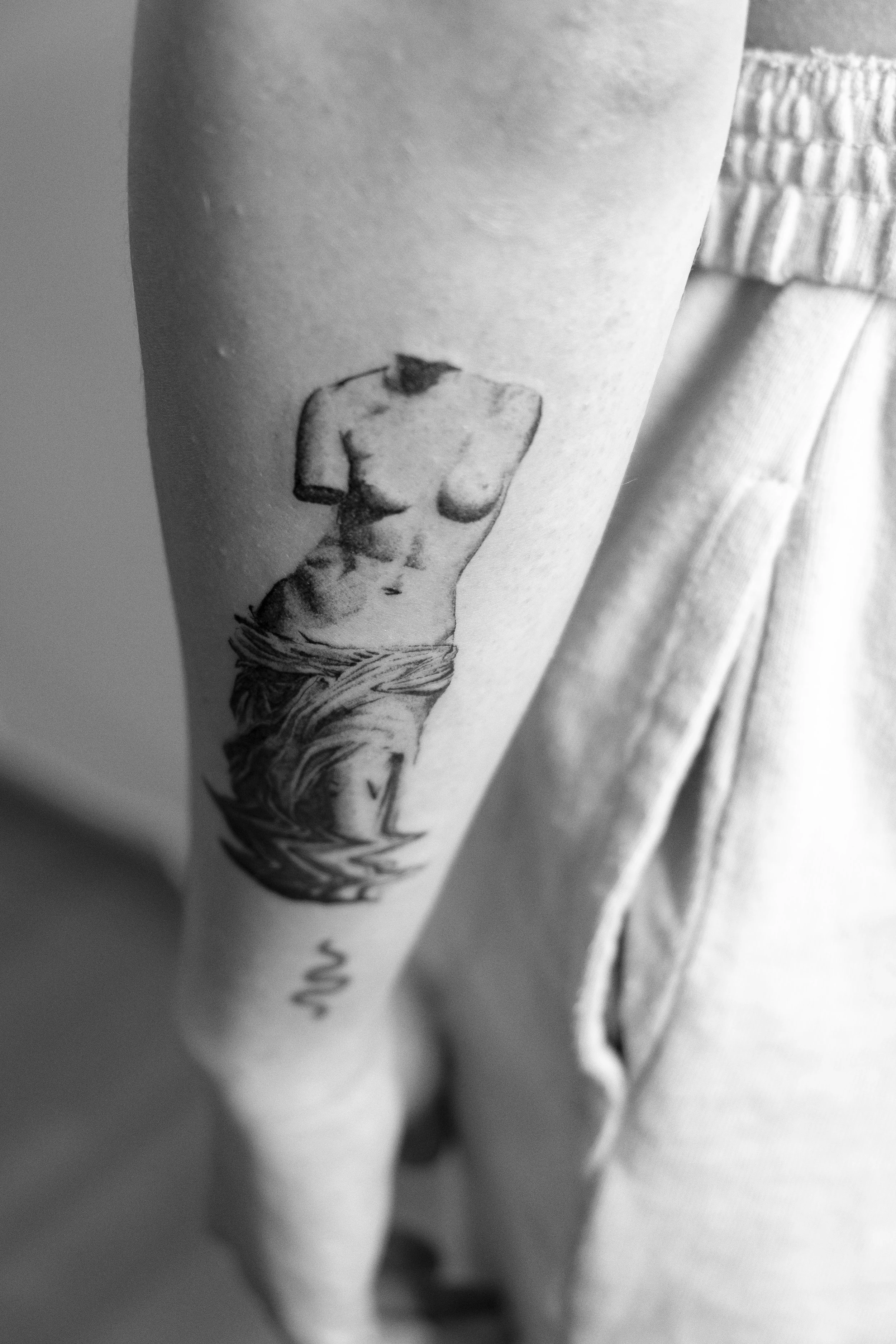 Schwarze Tätowierung einer weiblichen, halbnackten Statue auf dem Unterarm