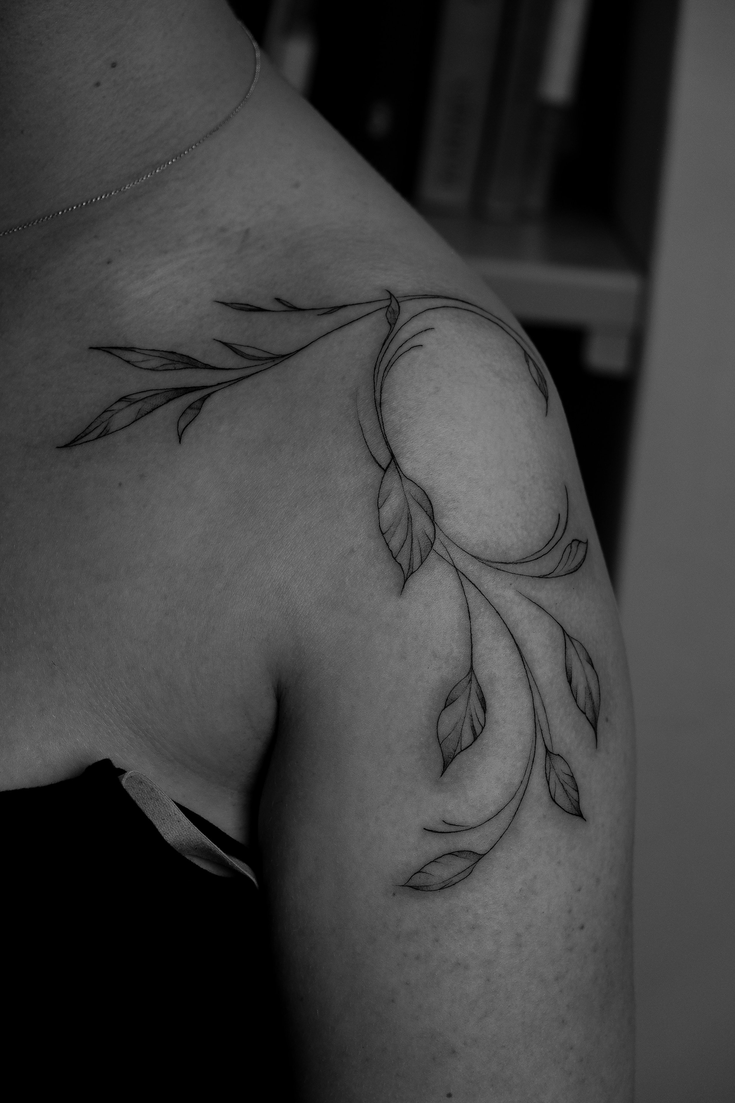 Schwarze und weiße Tätowierung eines Zweigs mit Blättern auf der Schulter eines Menschen.