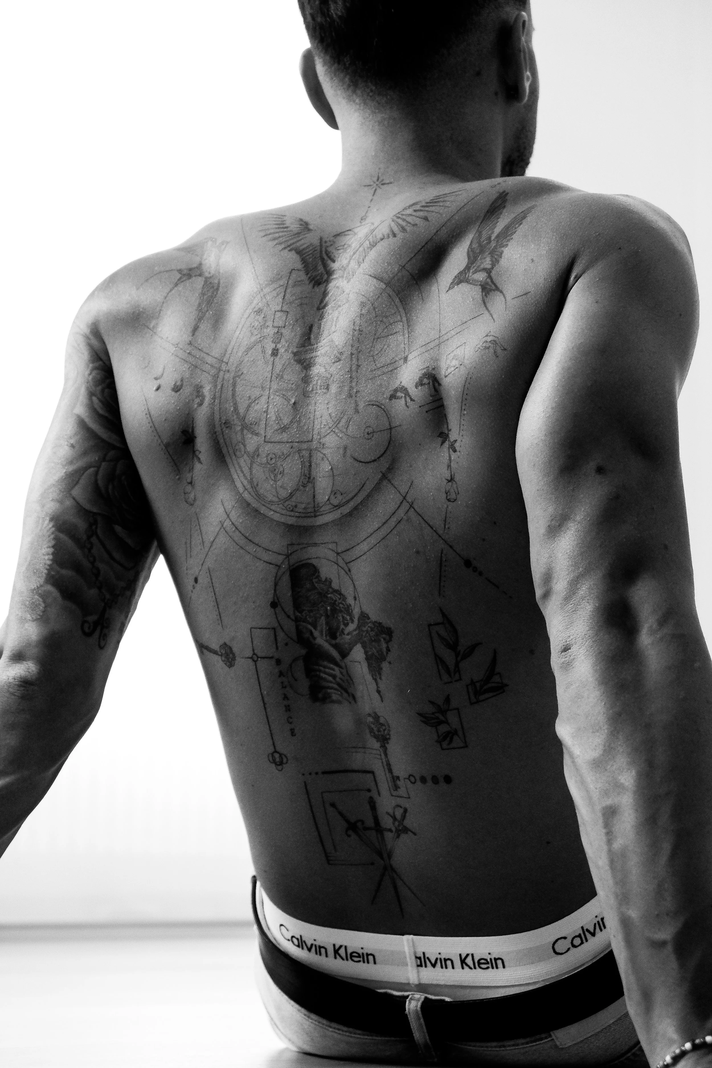 Schulter- und Rücken-Tattoo eines männlichen Models in Schwarz-Weiß-Fotografie, mit Calvin Klein Unterwäsche sichtbar.