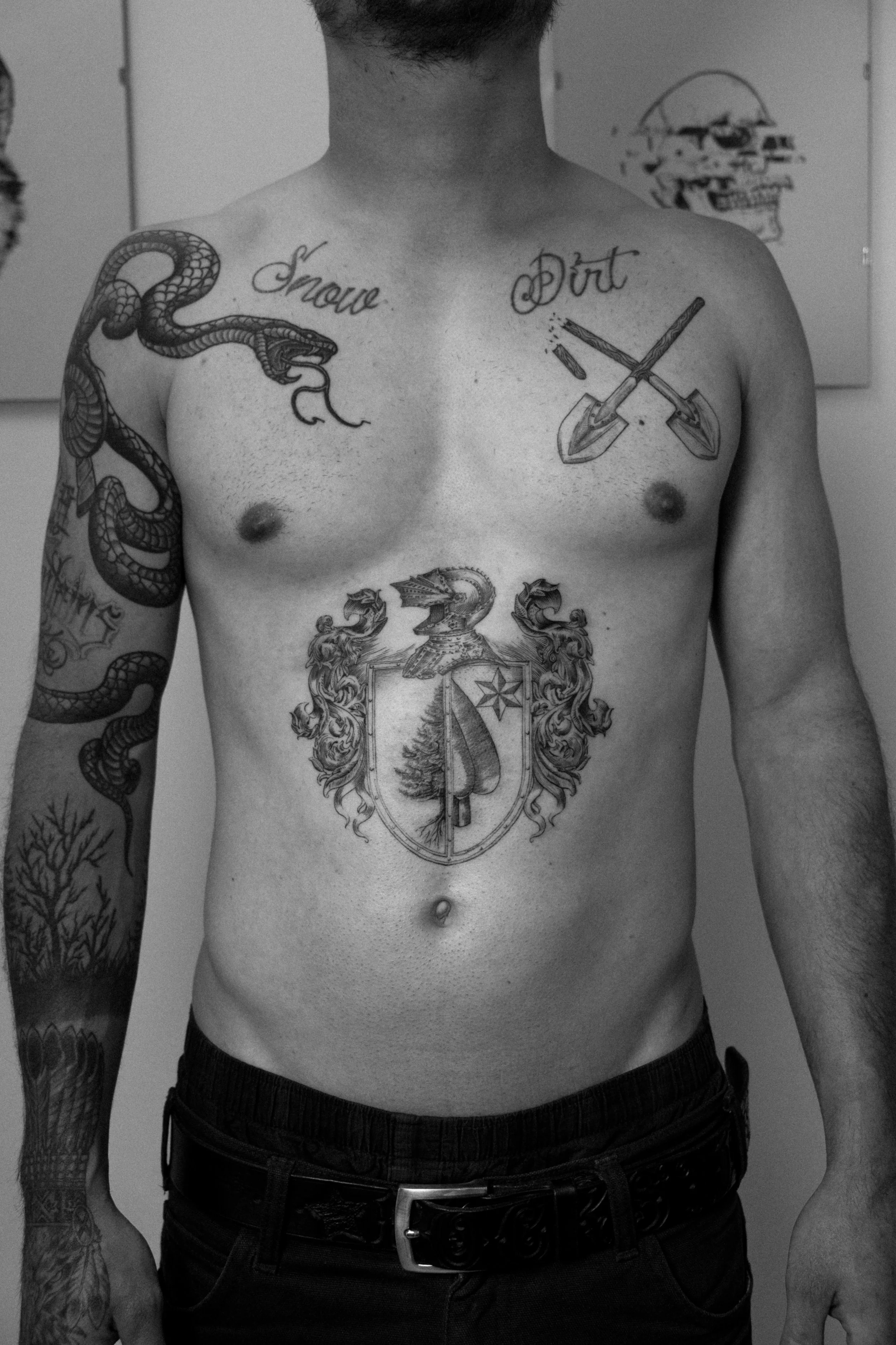 Mann mit Tattoos, darunter ein Schild mit einem Baum, ein Drache am rechten Arm, zwei Spaten auf der Brust, die Worte 'Snow' und 'Dirt' auf der Brust sowie ein Skull im Hintergrund.