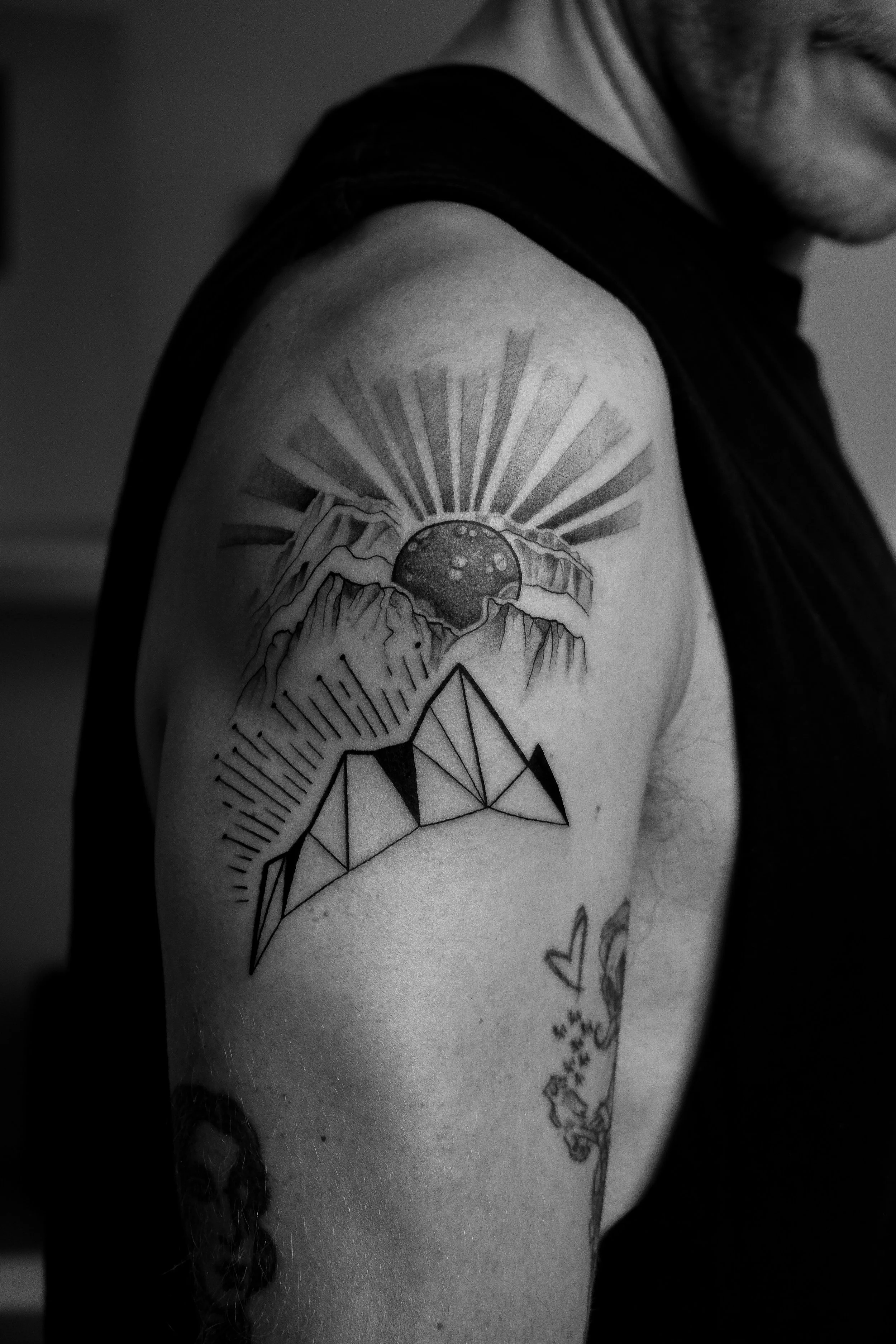 Schwarze und weiße Tätowierung auf einem männlichen Oberarm, zeigt eine bergige Landschaft mit Sonnenaufgang und geometrischen Mustern
