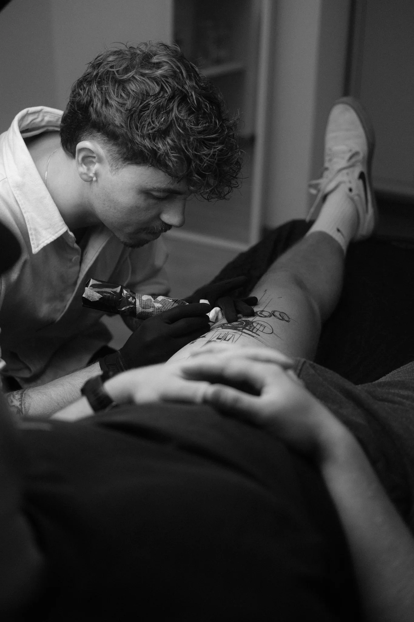 Ein Mann tätowiert die Wade eines anderen Mannes, der auf einer Liege liegt. Das Bild ist in Schwarzweiß.