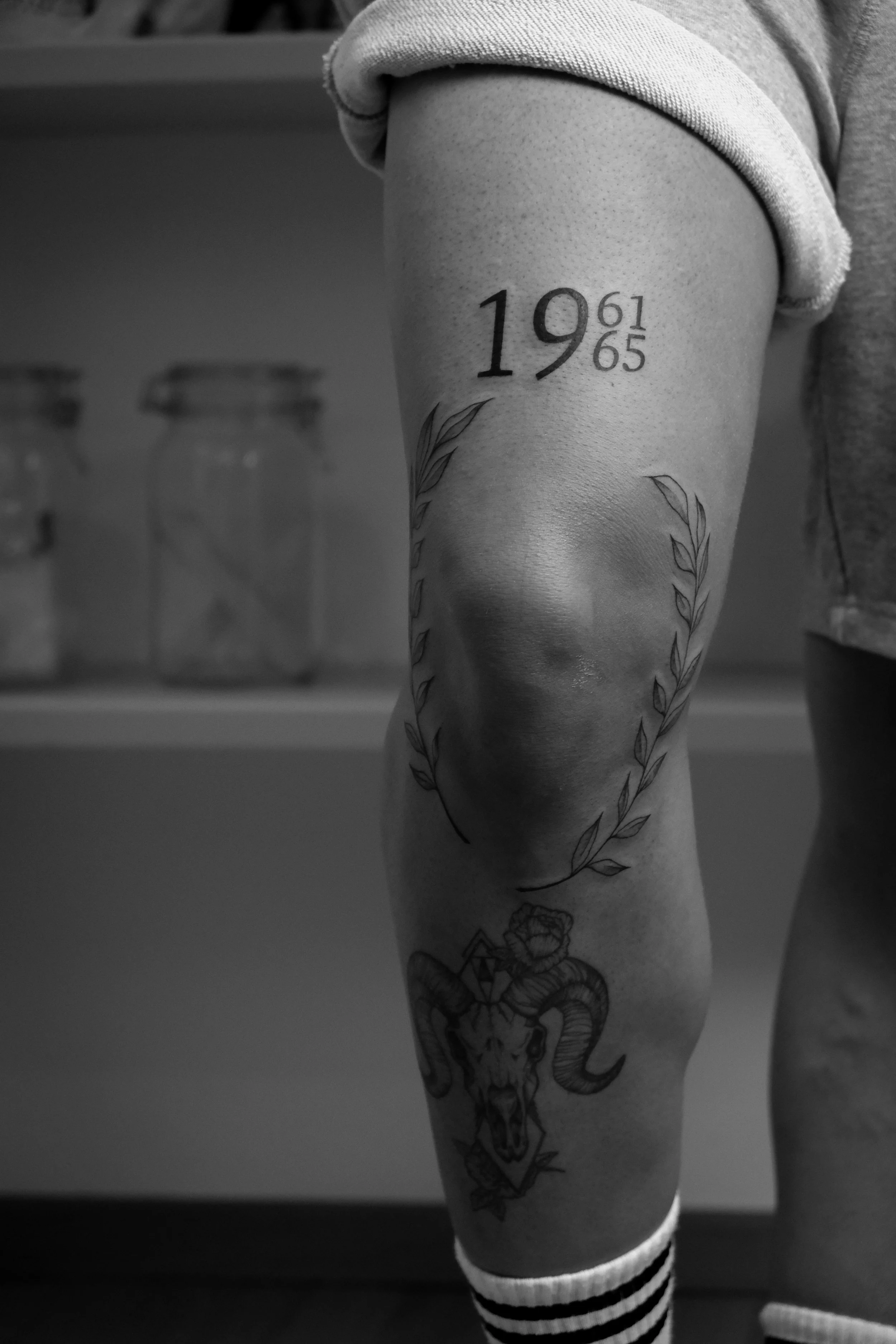Schwarze Tätowierung am Oberarm mit den Zahlen '19' und '65', umgeben von einem Lorbeerkranz, und unten ein Tierkopf mit Hörnern und einem Schädel, im Hintergrund sind Gläser im Kühlschrank sichtbar.