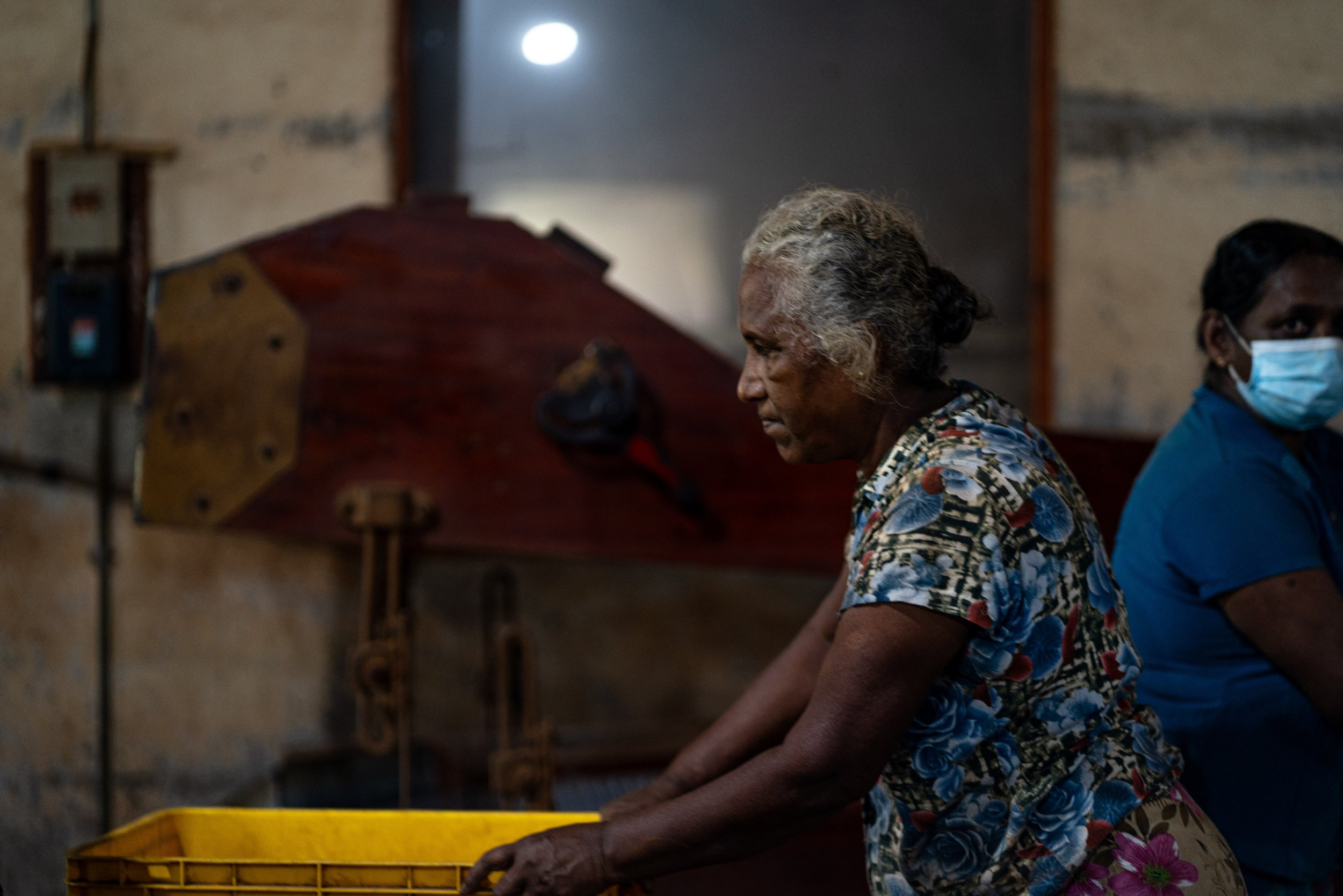 An elderly woman wearing a floral shirt pushes a yellow basket, with a woman wearing a blue shirt and face mask sitting beside her in a dimly lit room.