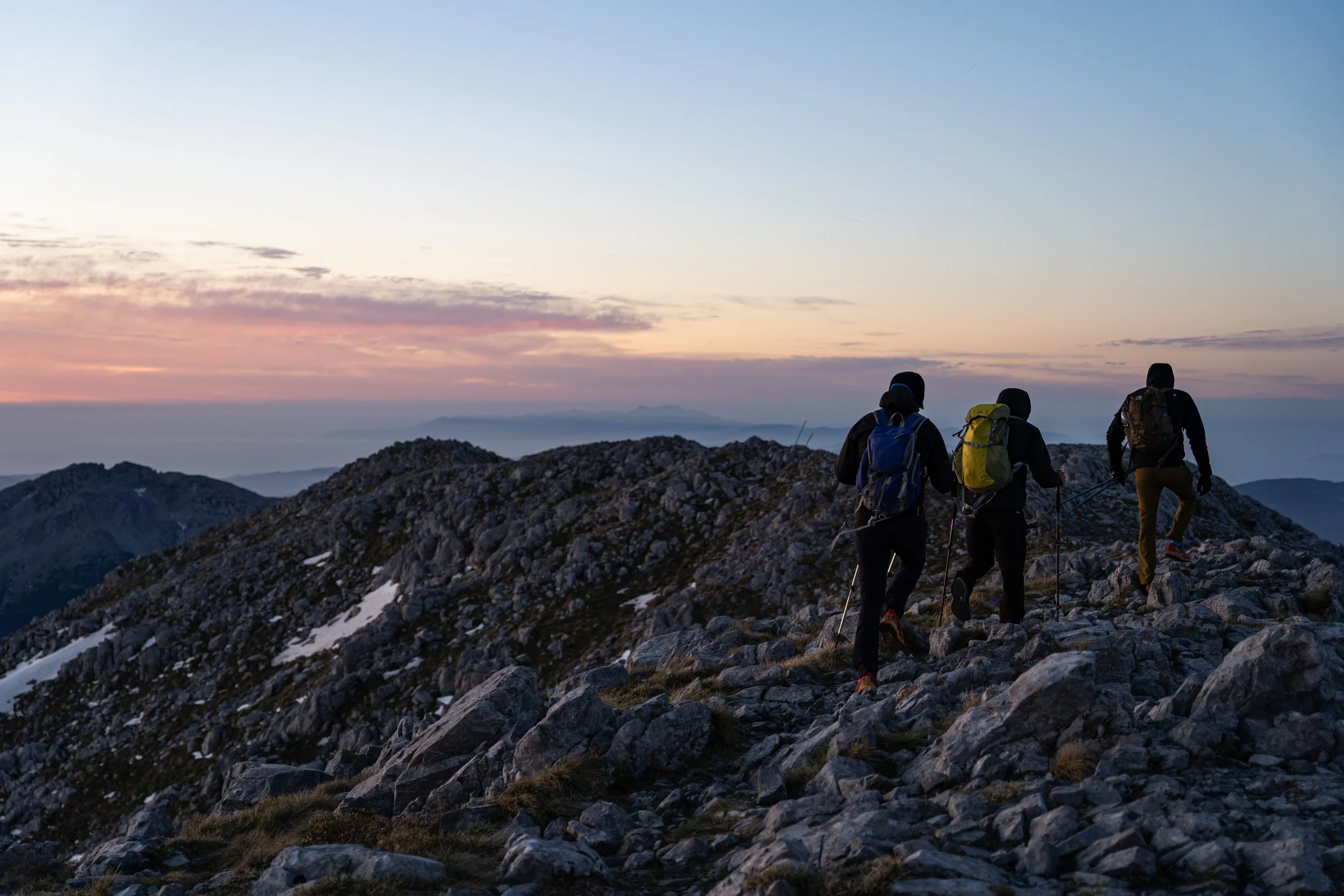 Four hikers with backpacks walking on a rocky mountain trail during sunset.