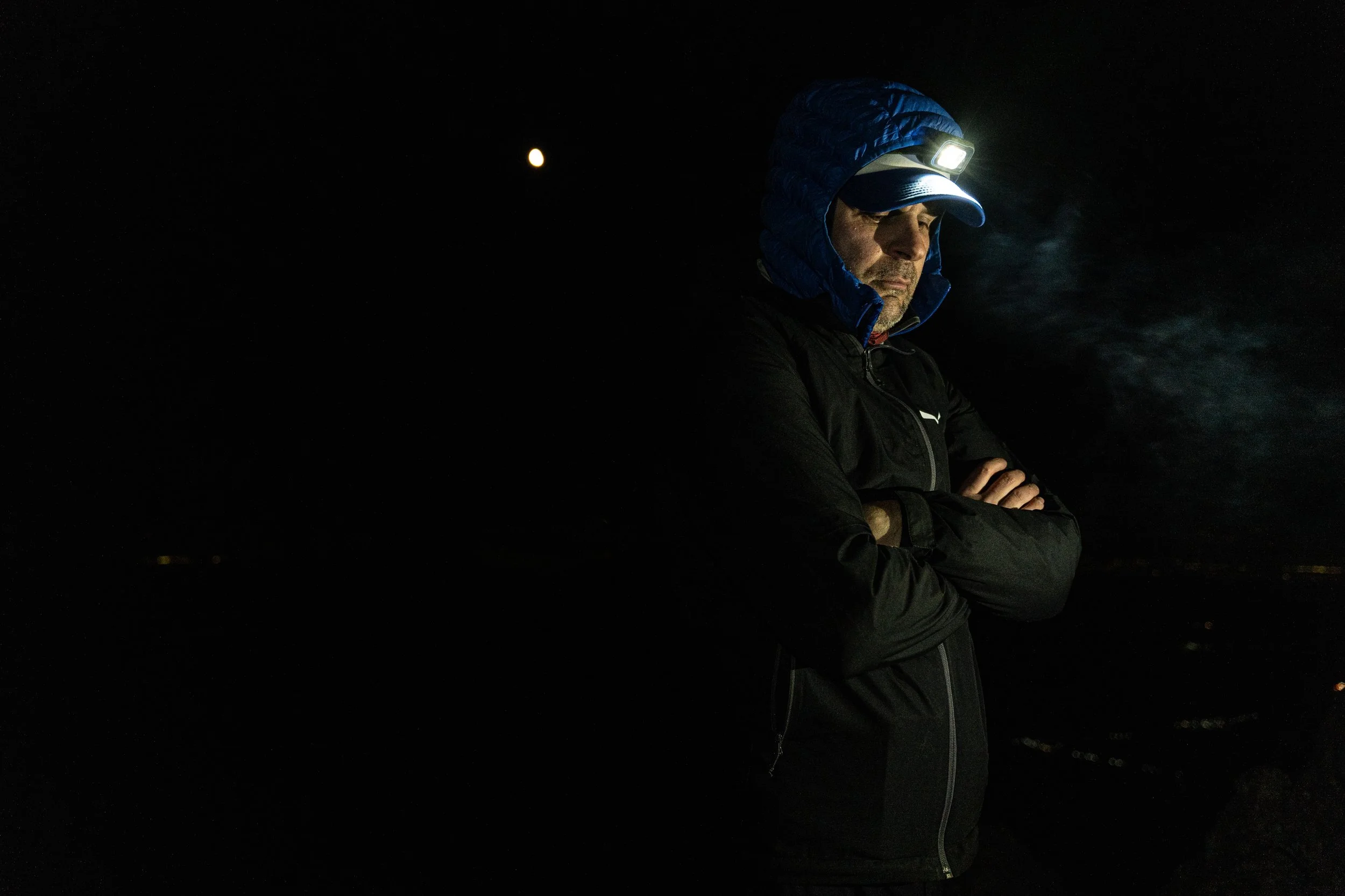 A man dressed in outdoor gear with a headlamp, standing in the dark outdoors with a visible moon in the sky.