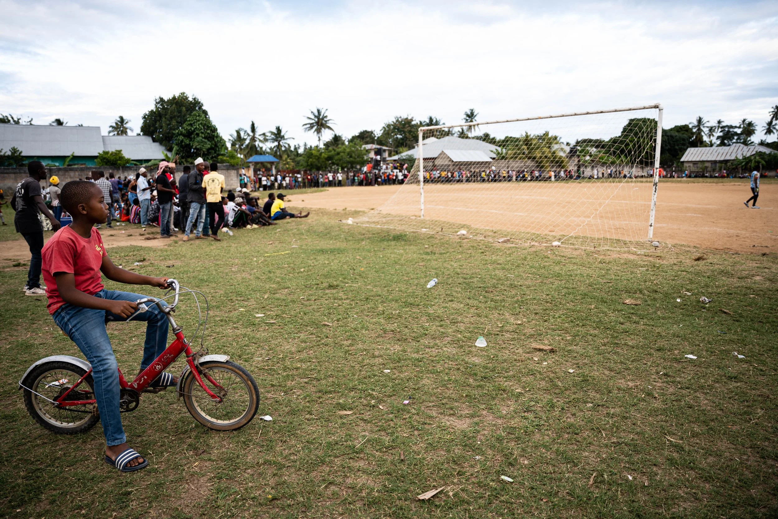 A young boy in a red shirt and jeans riding a red bicycle on a grassy field near a soccer goal, with a crowd of people gathered along the side of a dirt soccer field in the background, under a cloudy sky.