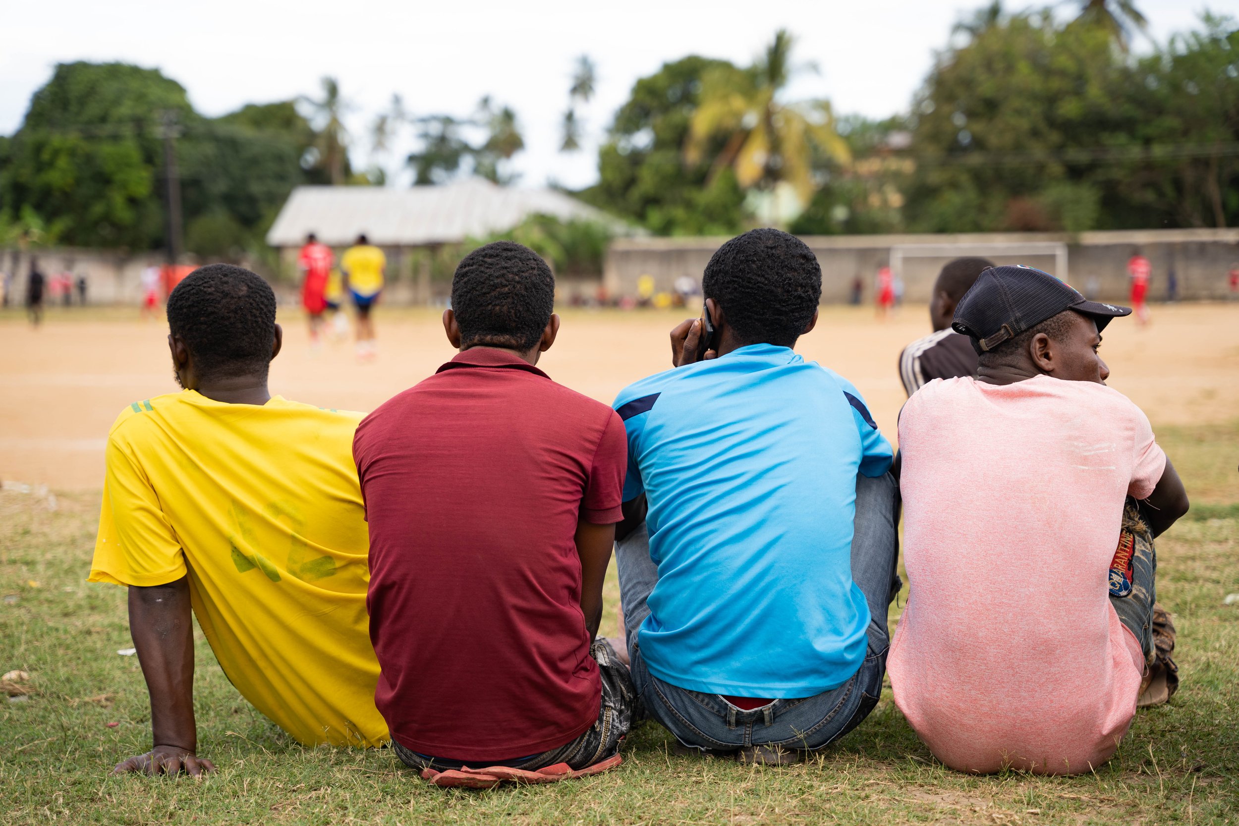 Four young men sitting on grass watching a soccer game on a field.