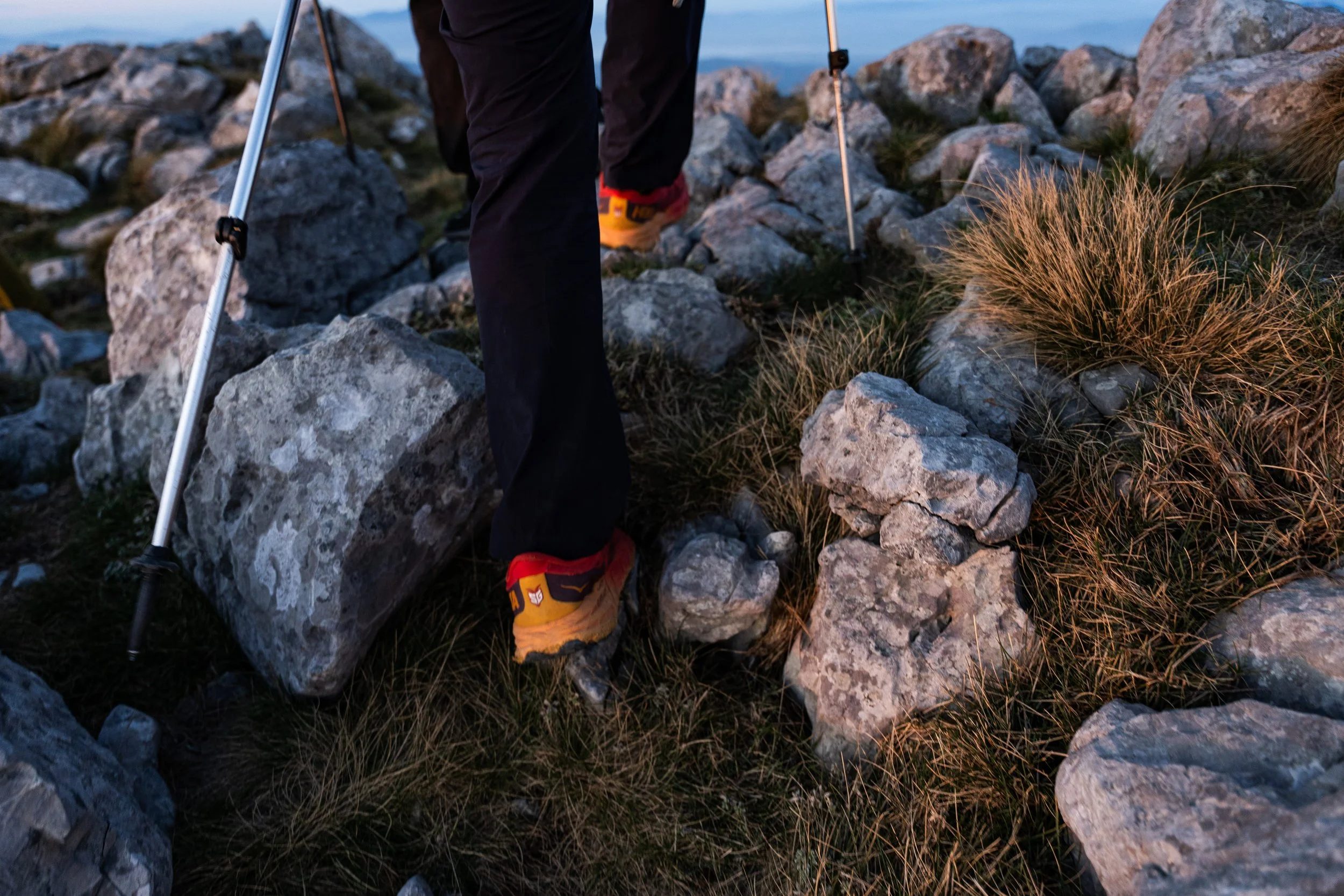 Close-up of a person hiking on a rocky trail, wearing orange and black hiking shoes, black pants, using trekking poles, in a mountainous terrain with rocks and sparse grass.