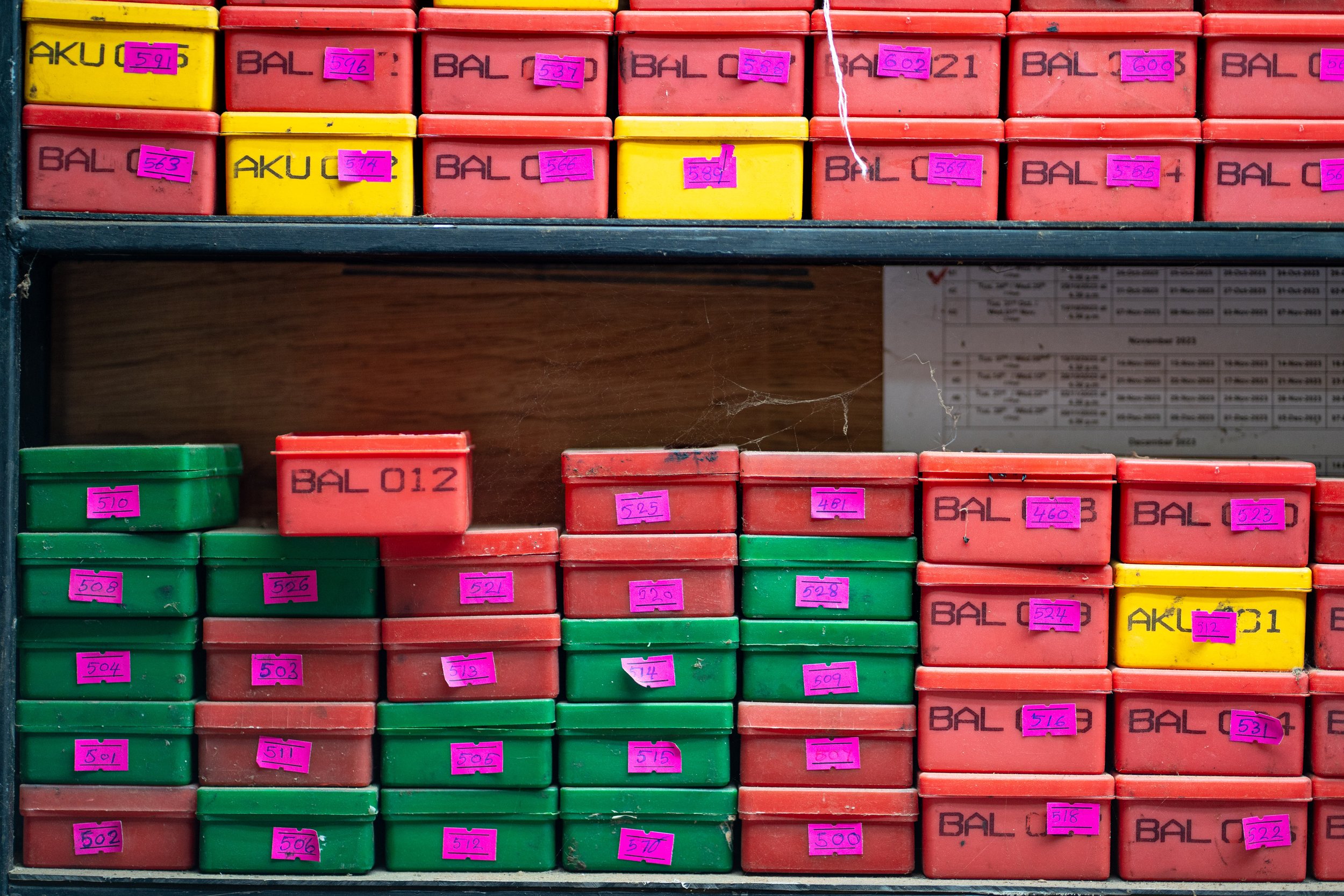 Shelf with multiple colorful boxes, mostly red and green, labeled with black text and pink stickers with handwritten numbers.