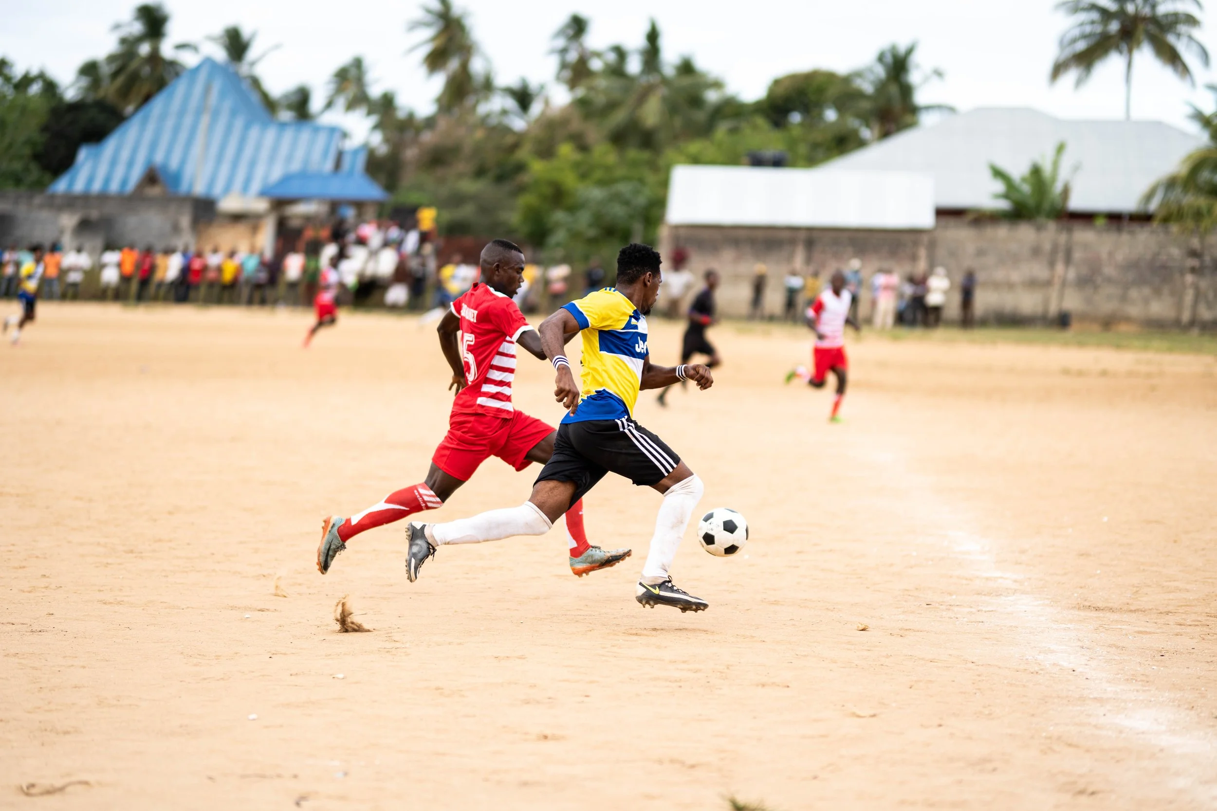 Two soccer players sprinting after the ball on a dirt field, with spectators and buildings in the background.