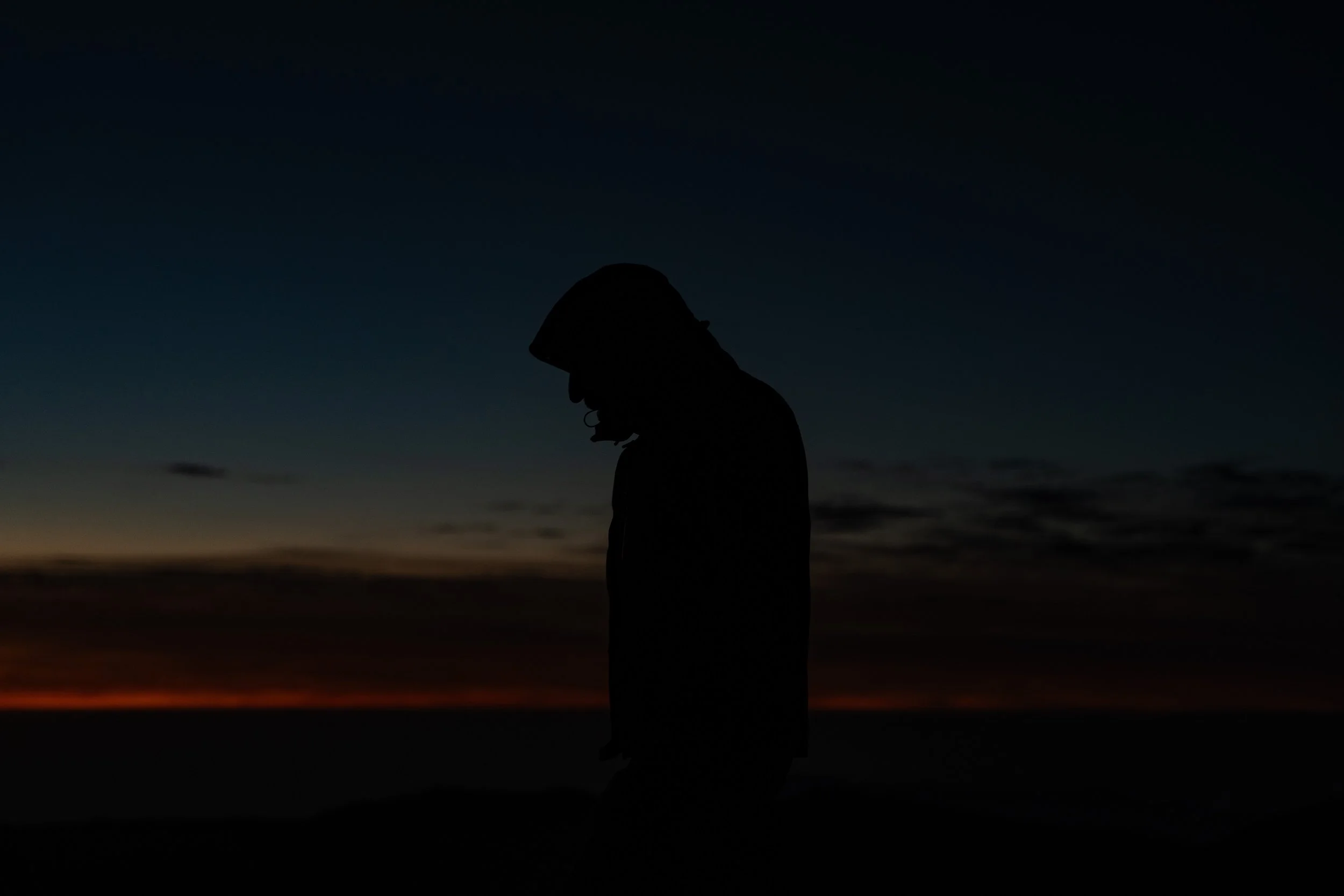 Silhouette of a person wearing a helmet, standing outdoors during sunset or sunrise with dark clouds on the horizon and a gradient sky transitioning from dark to light.