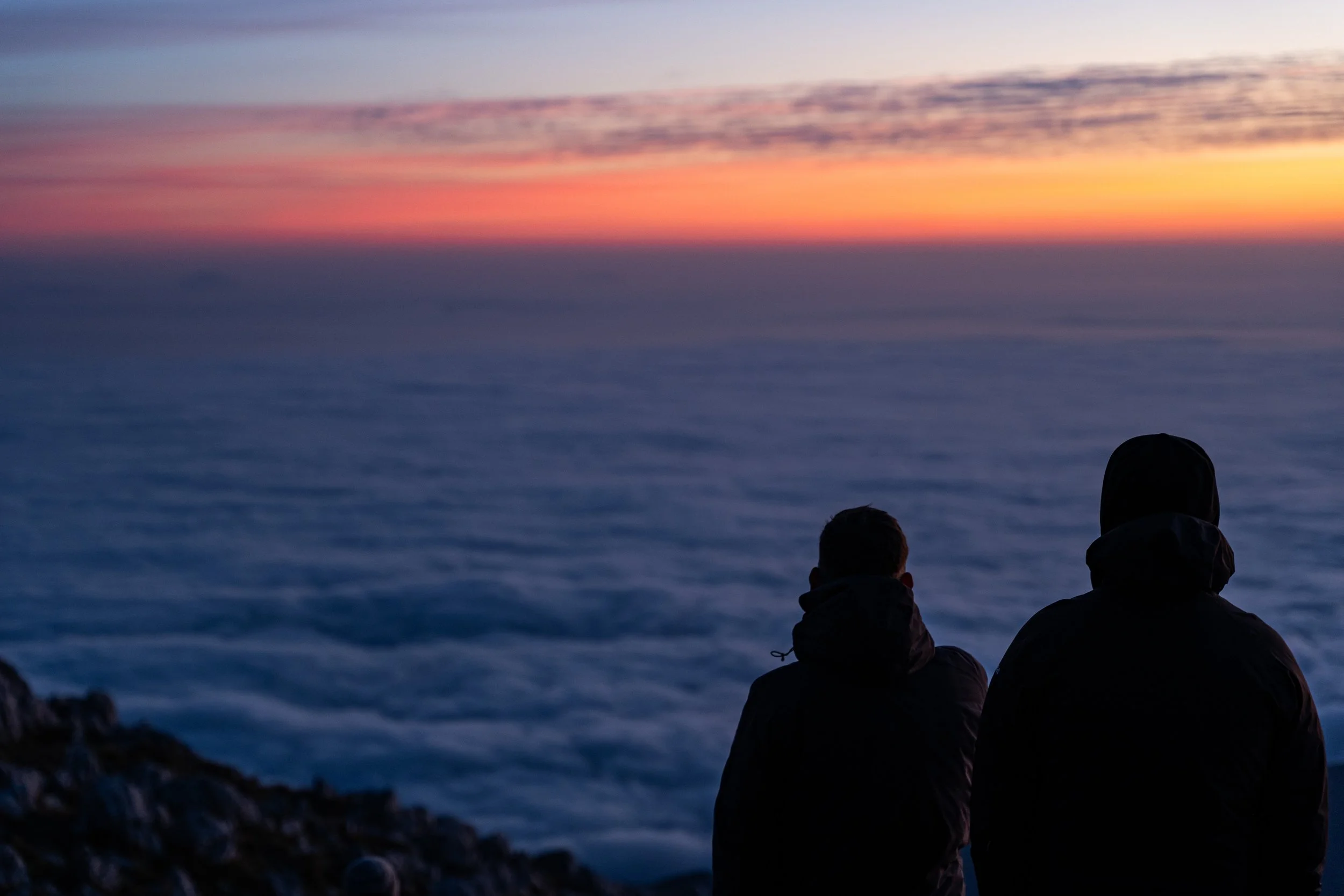 Two people wearing dark jackets sitting on a rocky ledge overlooking a body of water at sunset or sunrise, with a colorful sky and clouds reflecting on the water.