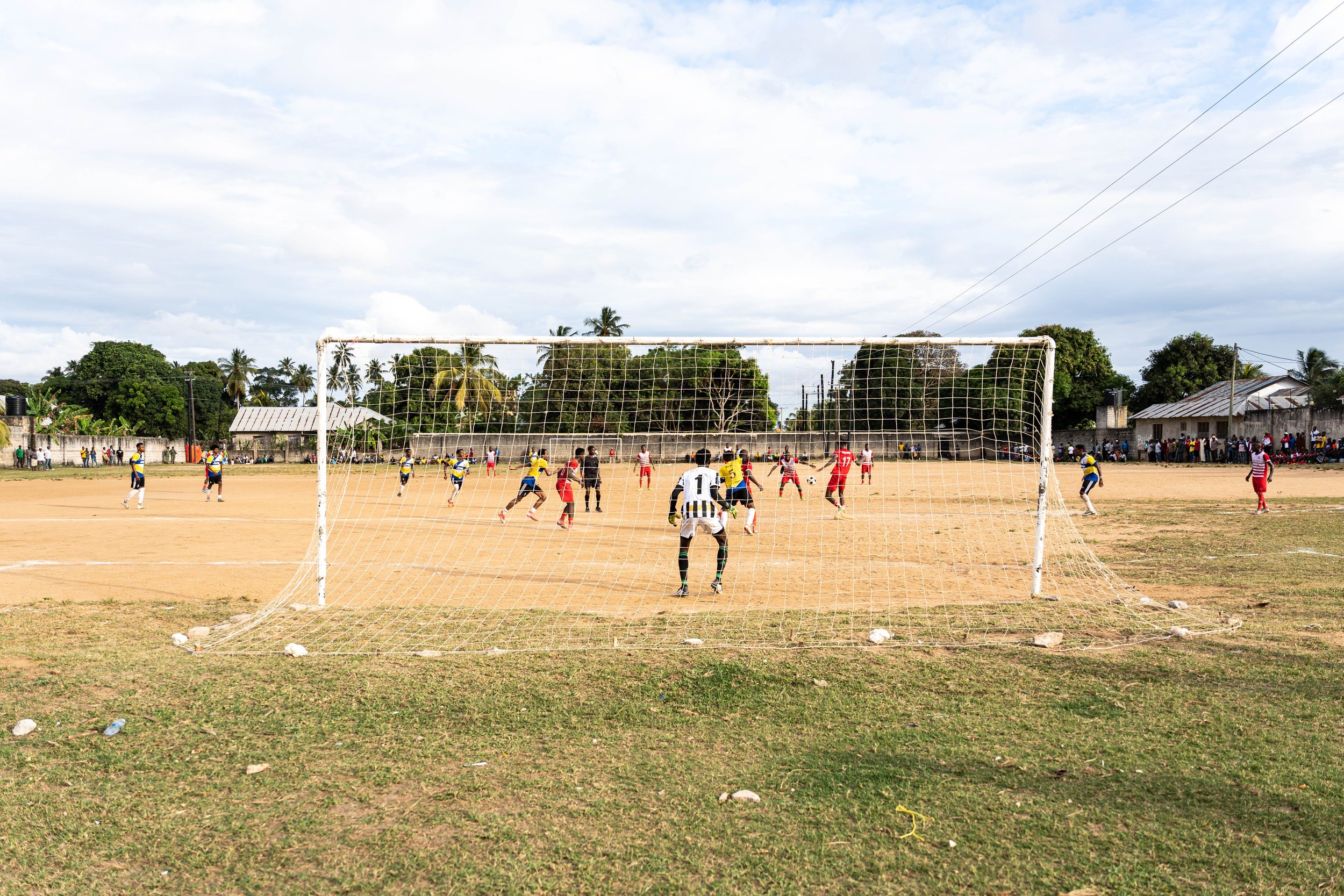 A soccer game in progress on a dirt field, with players wearing uniforms and a crowd watching from the sidelines and background. The goalpost is in the foreground.