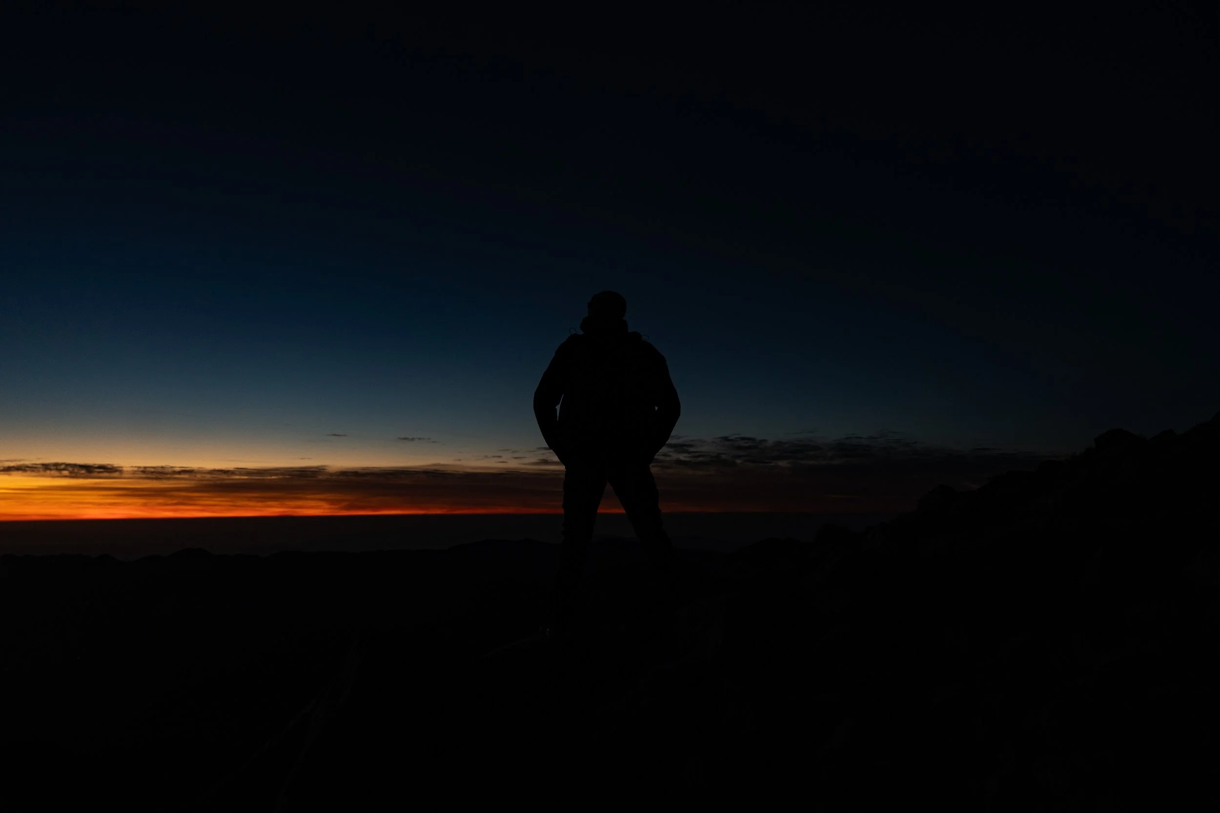 Silhouette of a person standing on a hill at sunset or sunrise, with a colorful sky and dark landscape.