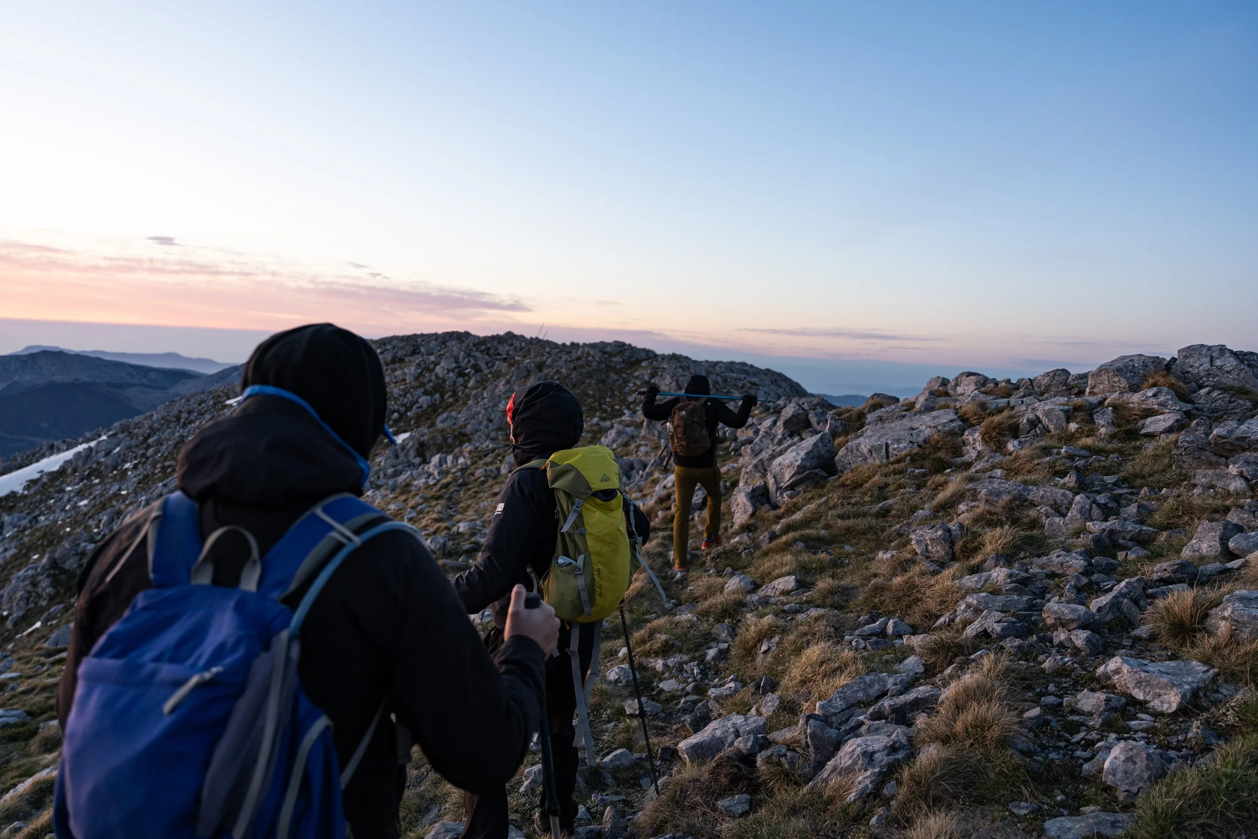 Three hikers in outdoor gear hiking on rocky terrain during sunset or sunrise in a mountain landscape.