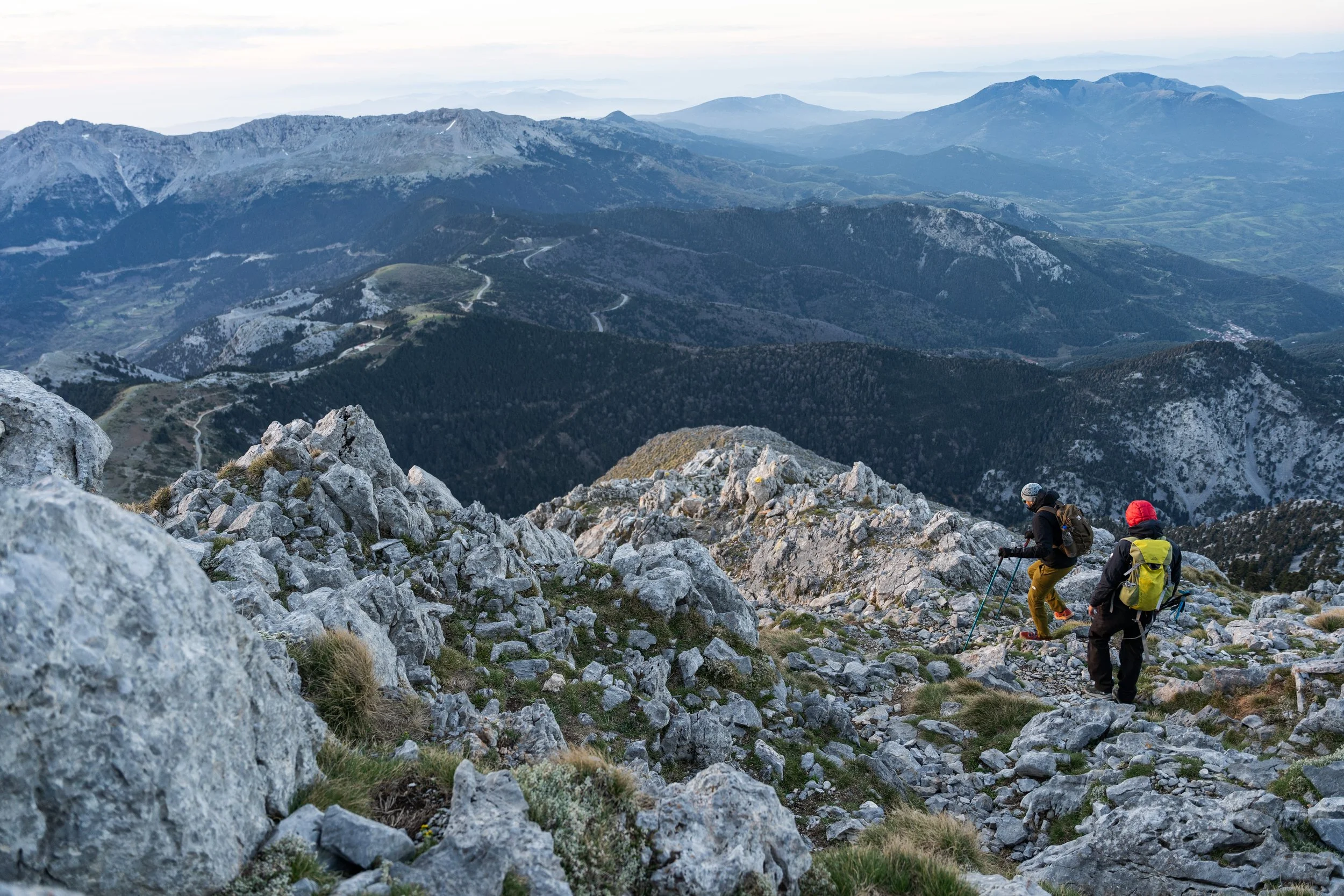 Two hikers ascending a rocky mountain trail with a vast mountain range in the background.