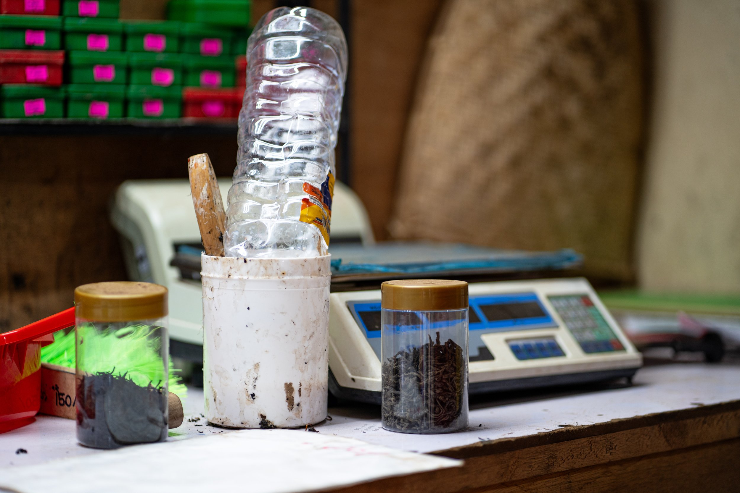 Desk with a plastic water bottle, two jars of dried tea leaves, a container with colorful feathers, a white cash register, and a digital scale in the background.