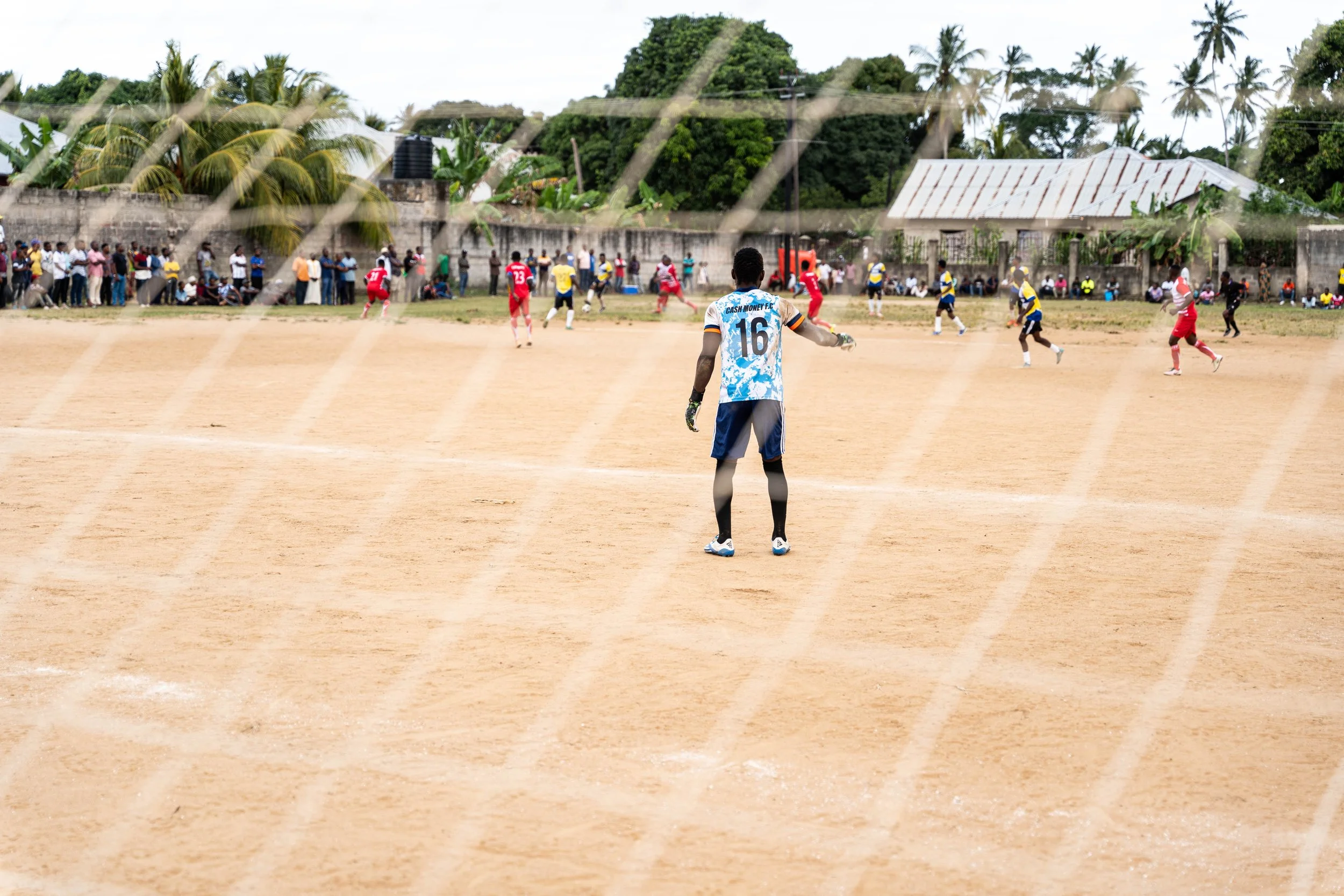 A soccer match on a dirt field viewed through a chain-link fence with a goalie in the foreground and players in red and blue jerseys in the background, with spectators watching from the sidelines.