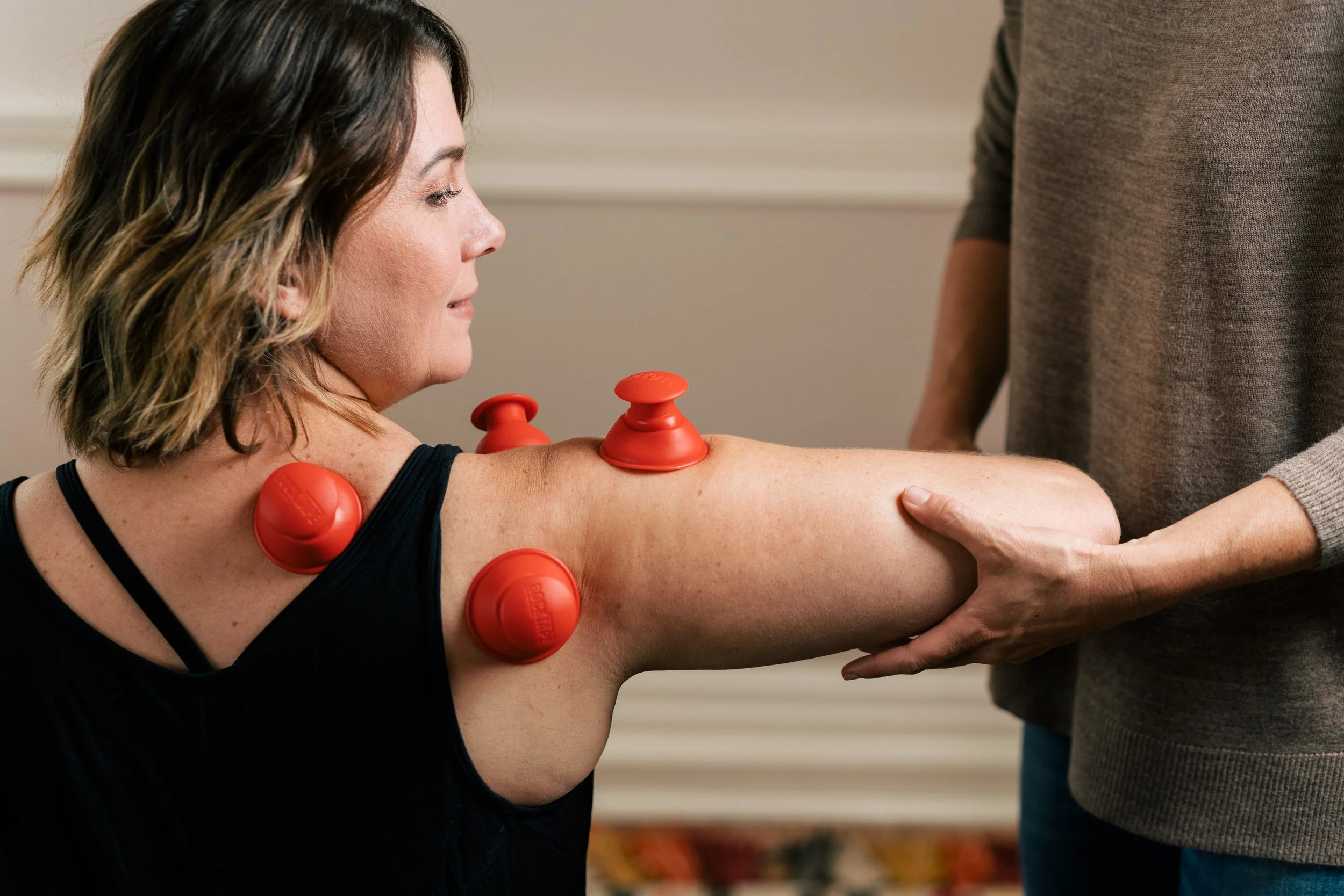 A woman with shoulder-length brown hair receiving physical therapy with red massage cups on her arm, while a therapist holds her arm and a third person stands nearby.
