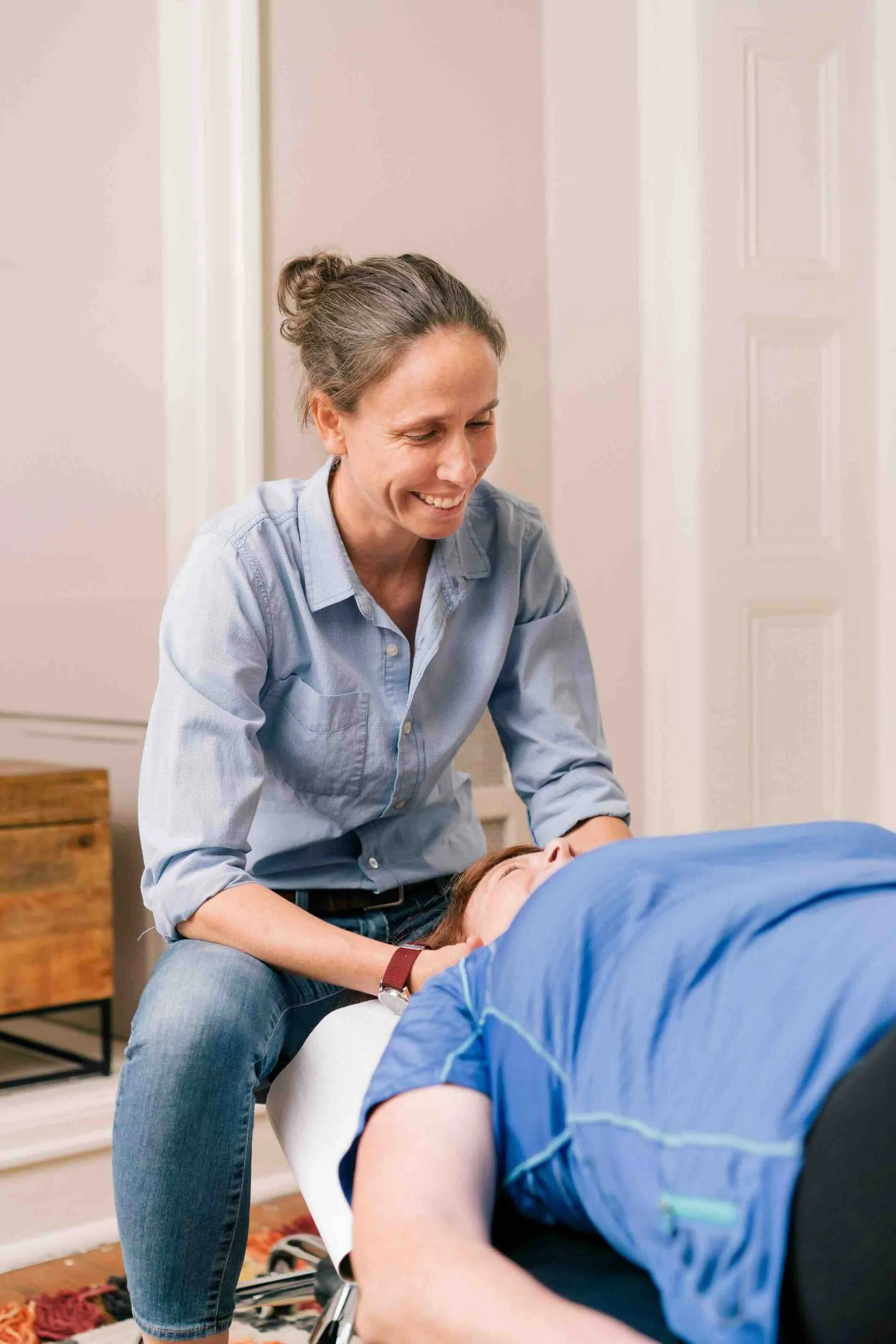 A woman with gray hair in a bun providing chiropractic care to a man lying face up on an exam table, with a smile on her face. The man is wearing a blue shirt and is being treated in a cozy, well-lit room with a wooden floor and a colorful rug.