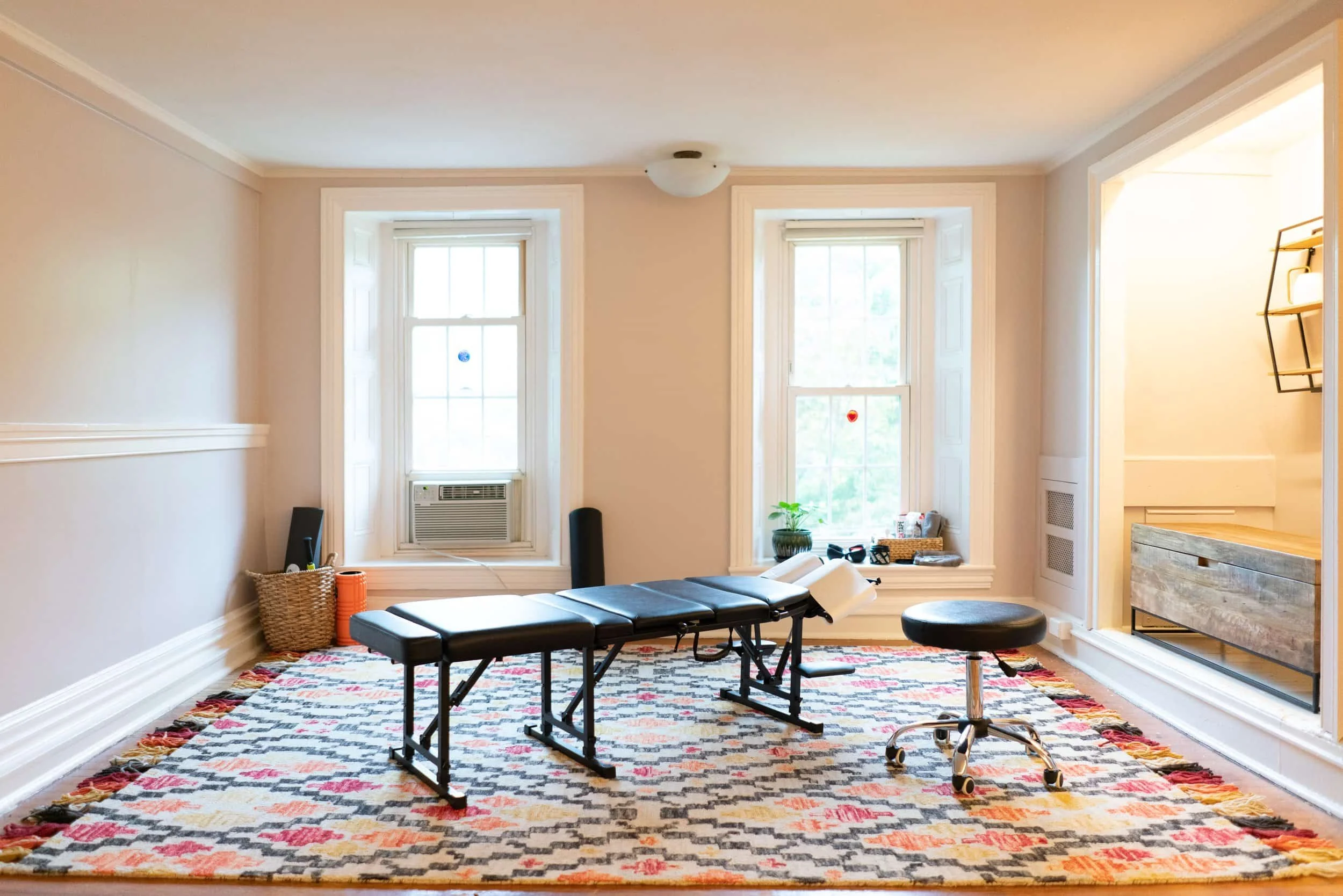 A living room with two large windows, an air conditioning unit, a colorful patterned rug, a black massage table, a rolling chair, a small basket with items, and a wall with a shelf in the corner.