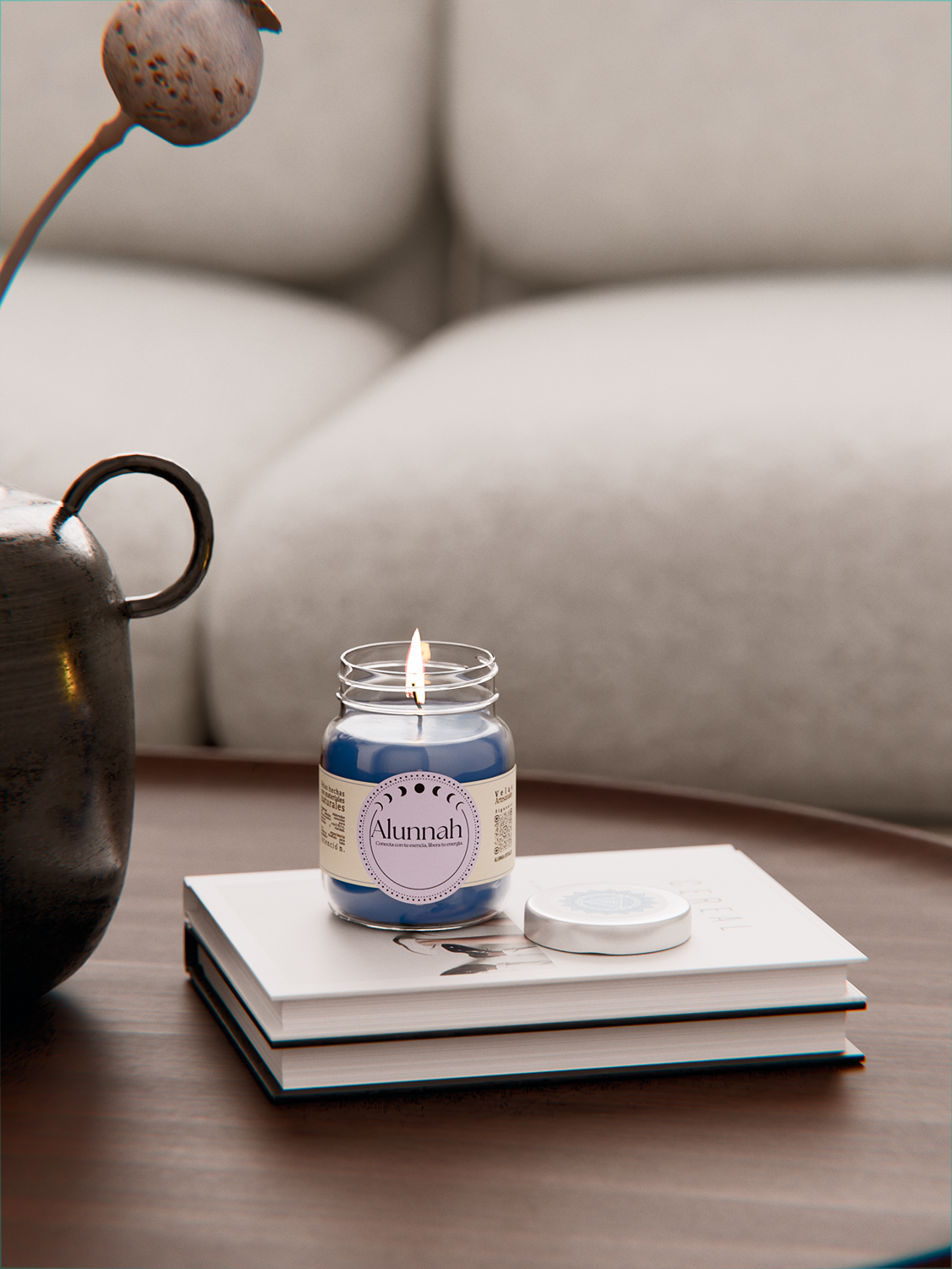 A lit blue candle labeled 'Alunnah' placed on top of a closed book, with a white container on the book, on a wooden table next to a decorative metal teapot. In the background, a beige upholstered sofa is visible.