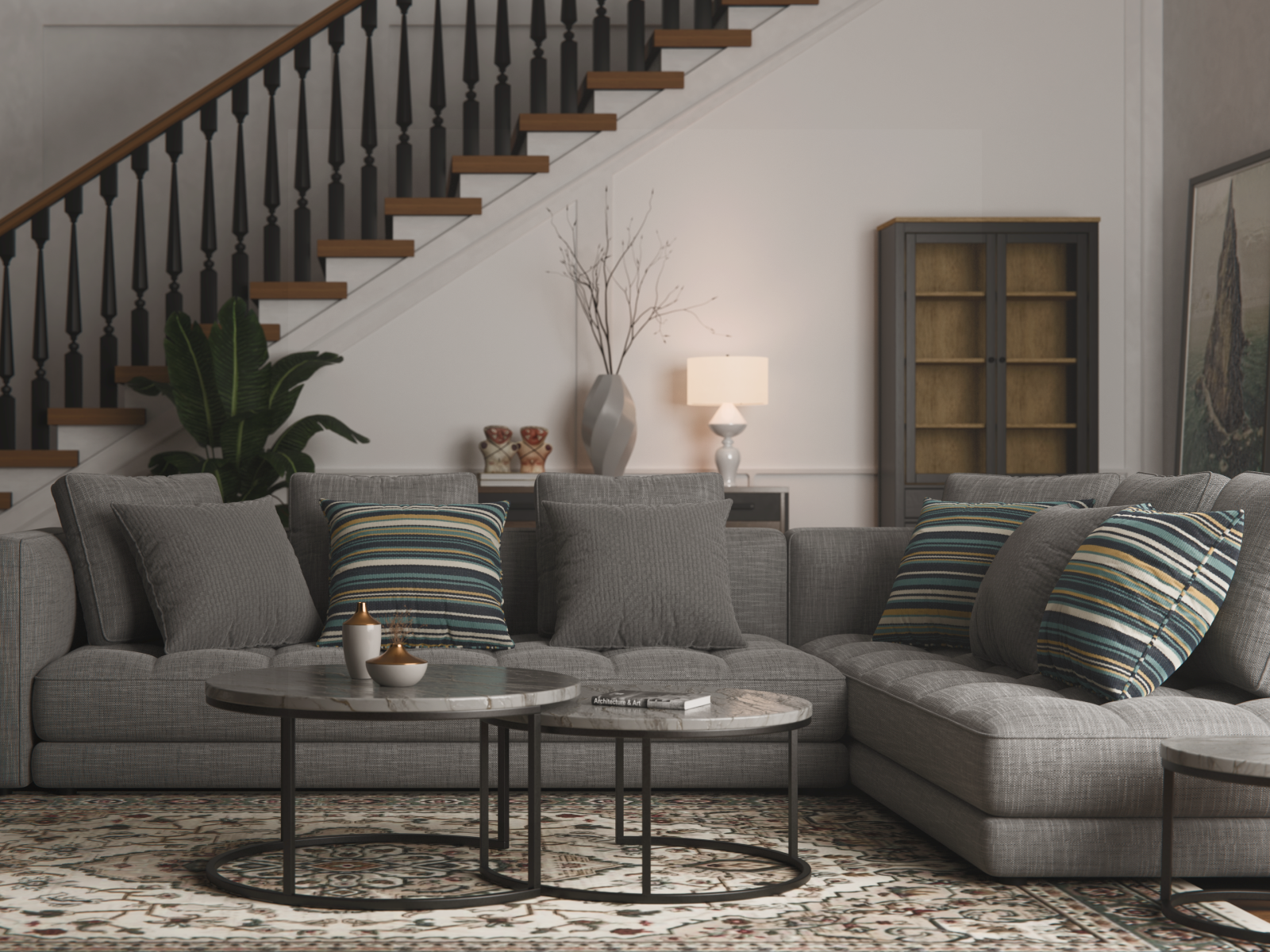 Living room with gray sectional sofa, striped and gray pillows, two round coffee tables, a large plant, a tall vase with branches, a table lamp, a cabinet, and a staircase in the background.