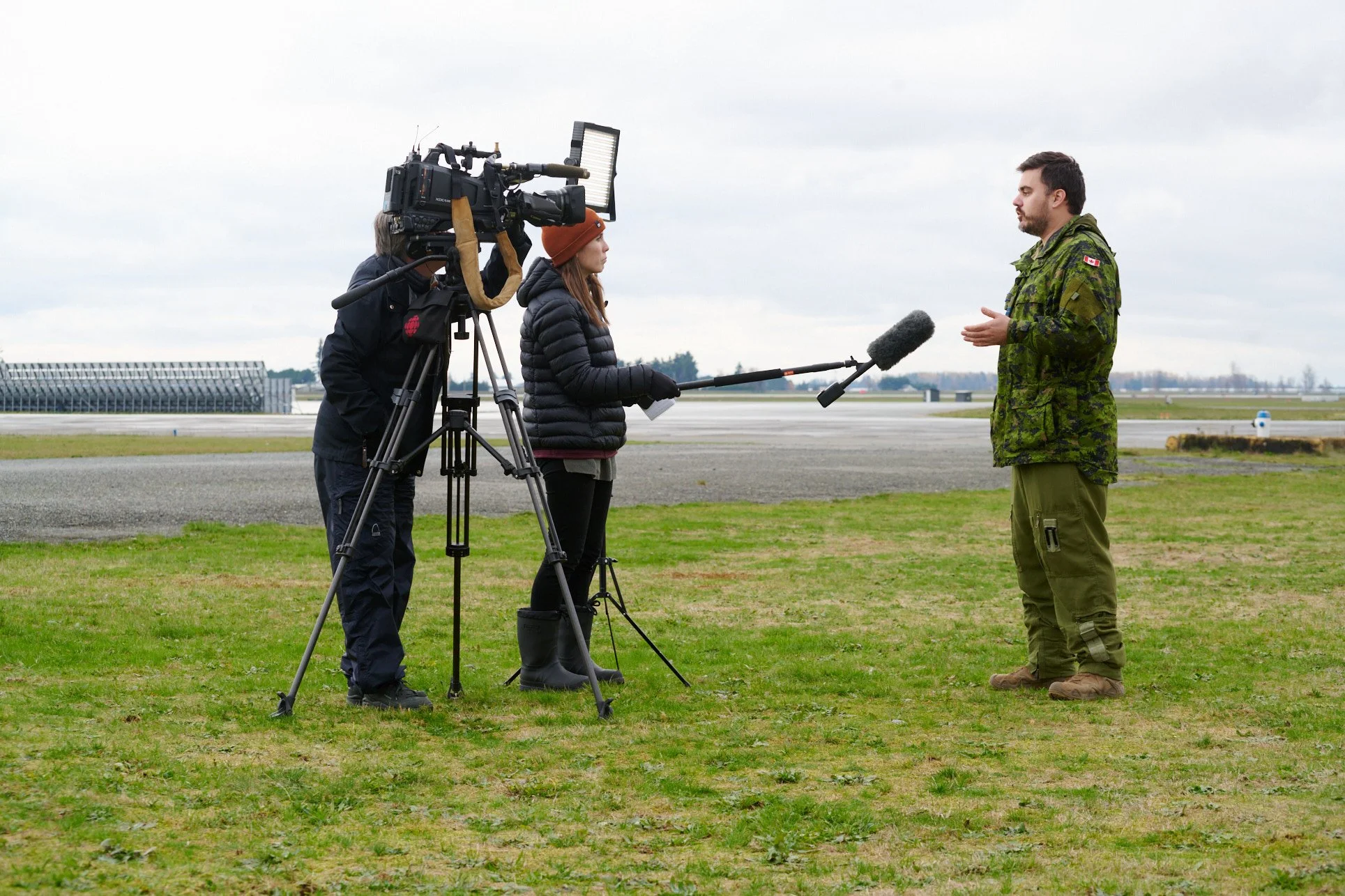 A news crew is filming an interview with a soldier at an airport tarmac on a cloudy day. The crew includes a woman reporter holding a microphone boom, a cameraman operating a camera, and a man in military uniform speaking to the camera. The backgroun