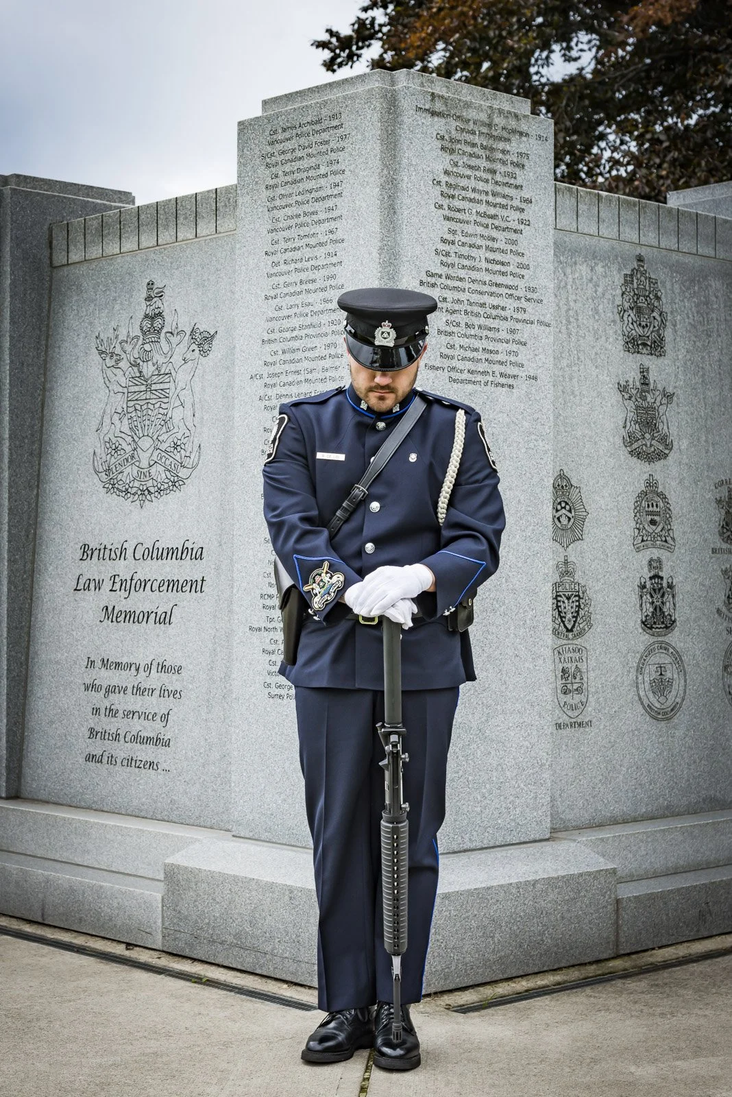 A police officer in a formal uniform standing at attention with a rifle in front of a memorial wall with engraved text and crests, dedicated to British Columbia law enforcement.