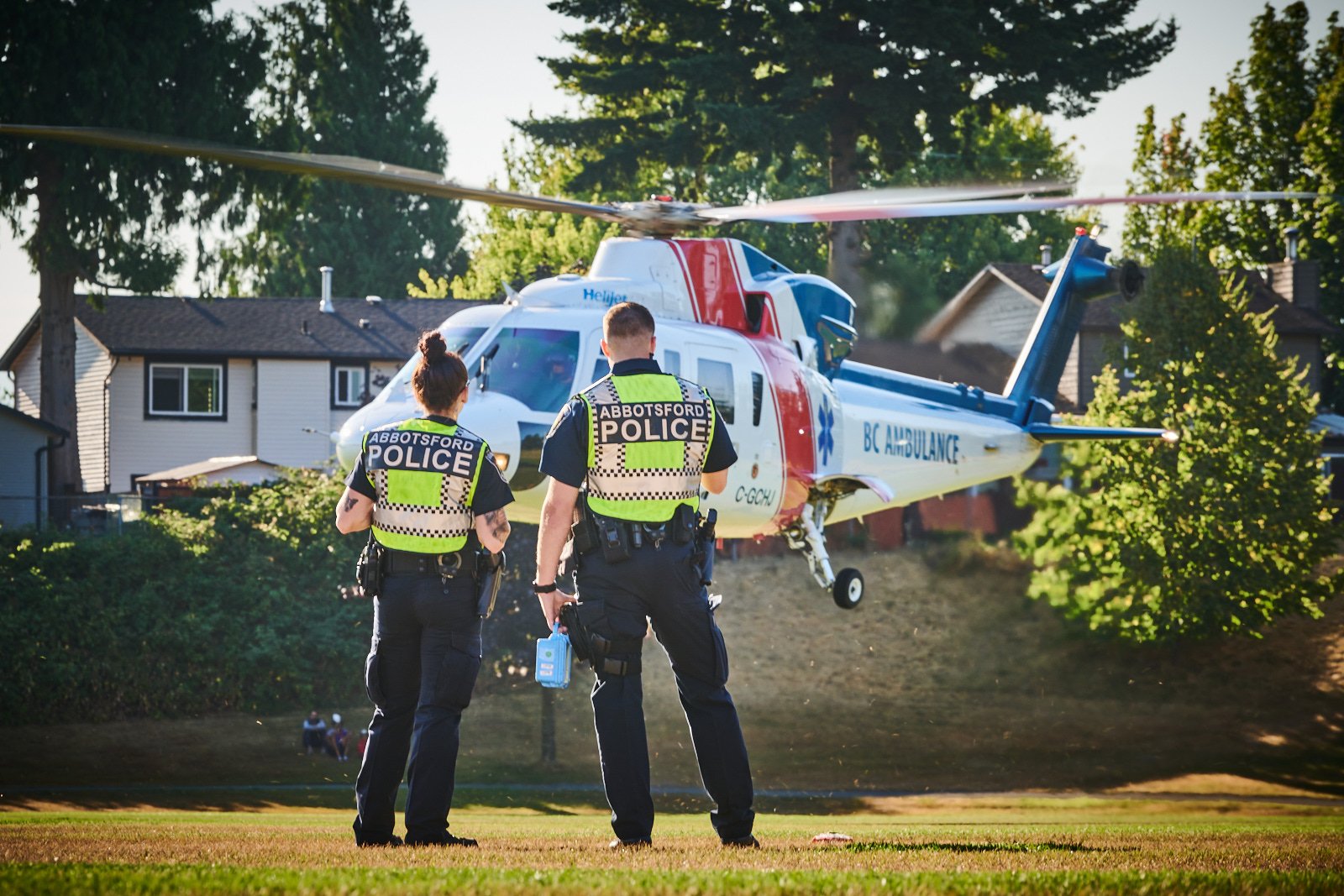Two police officers in reflective vests standing on grass near a BC Ambulance helicopter, which is hovering above a suburban area with houses and trees.