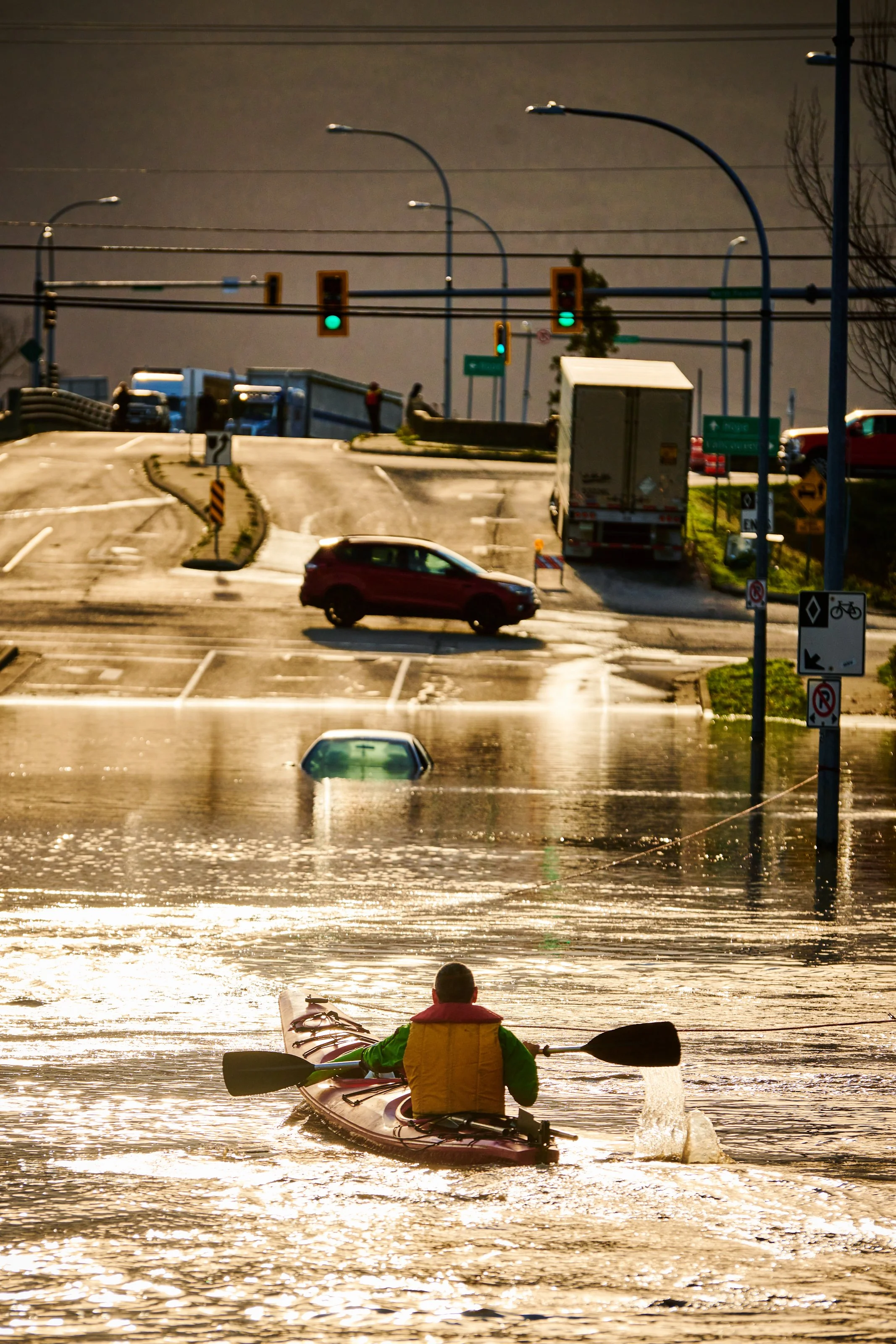 A person in a yellow life jacket paddling a kayak in floodwaters at sunset, with submerged road and cars in the background.