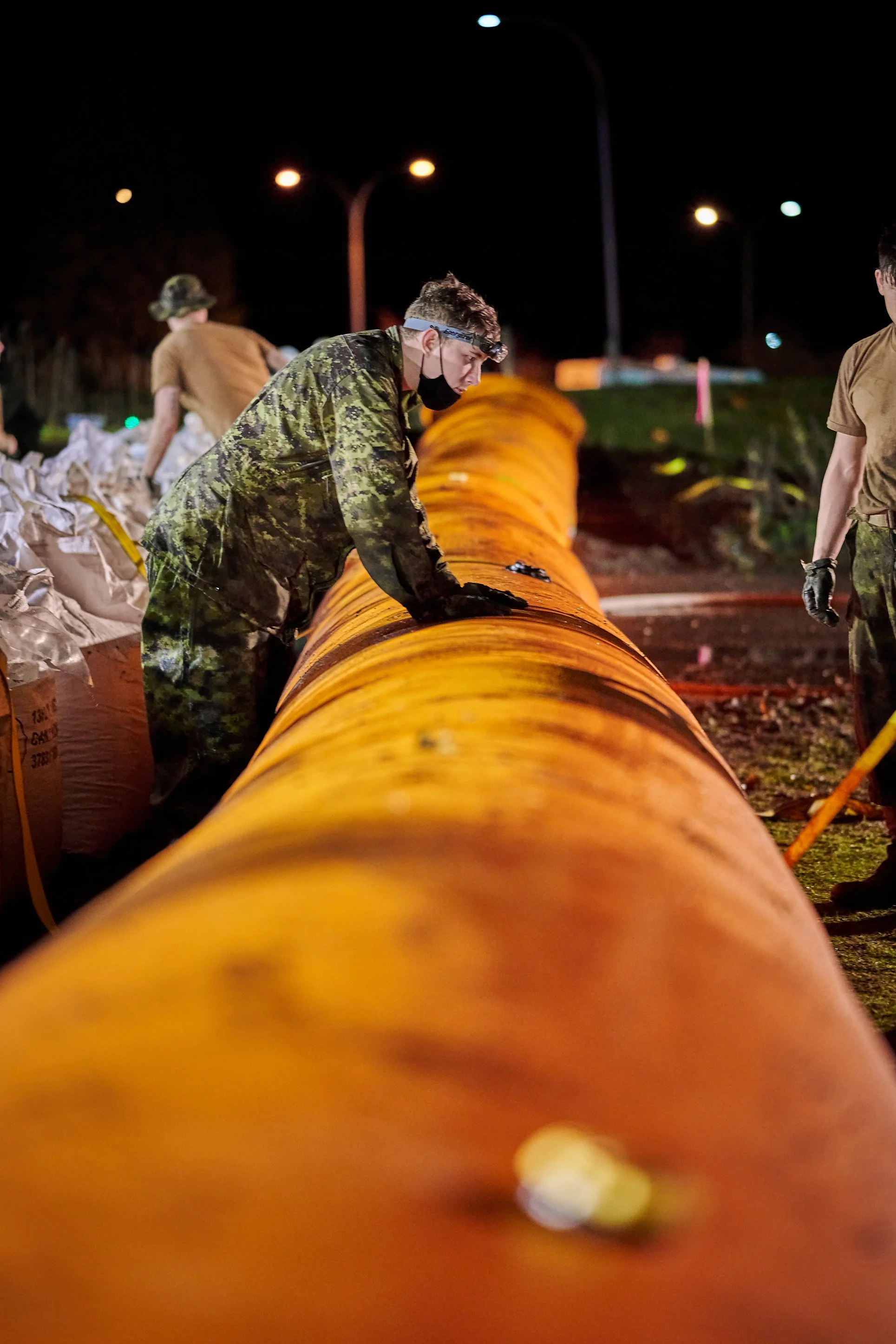 Nighttime scene of people working on a large orange pipe outdoors, with some wearing camouflage uniforms and headlamps.