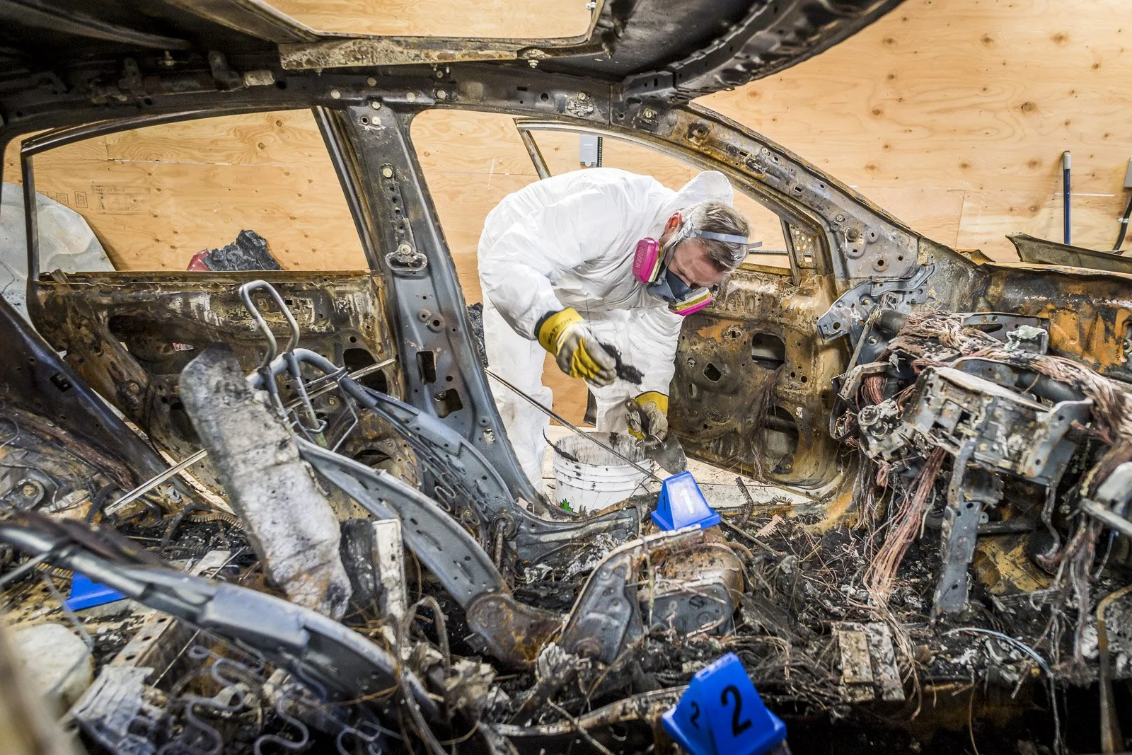 A person wearing a white protective suit, gloves, goggles, and a respirator mask is working with tools inside a burnt, disassembled vehicle in a workshop or forensic lab.