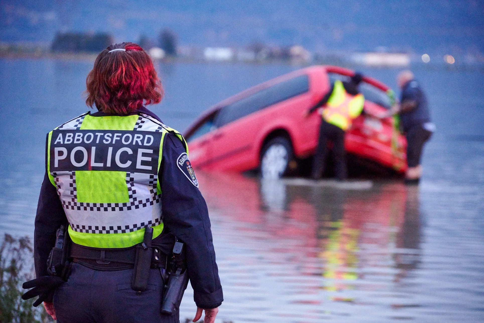 A police officer from Abbotsford is standing near a body of water, observing a red car that is partially submerged and tilted in the water. Two other officers are attending to the vehicle in the background.