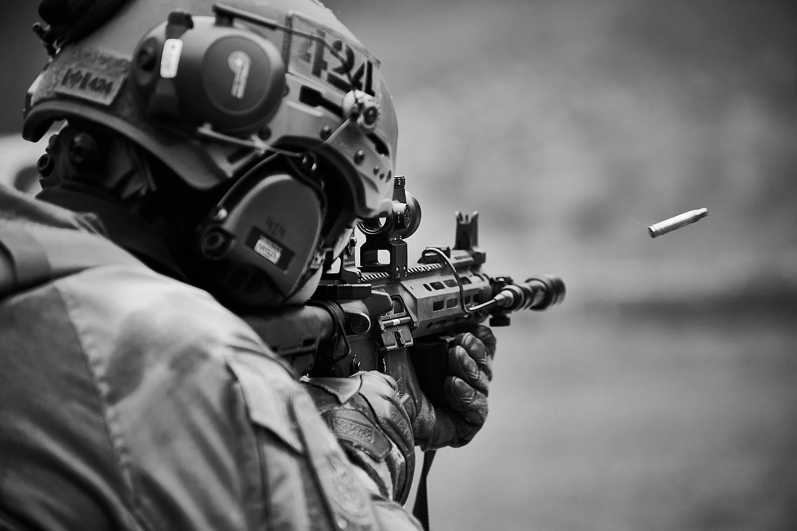 A soldier aiming a rifle with a bullet in mid-air, wearing a helmet and tactical gear, in a black and white photograph.