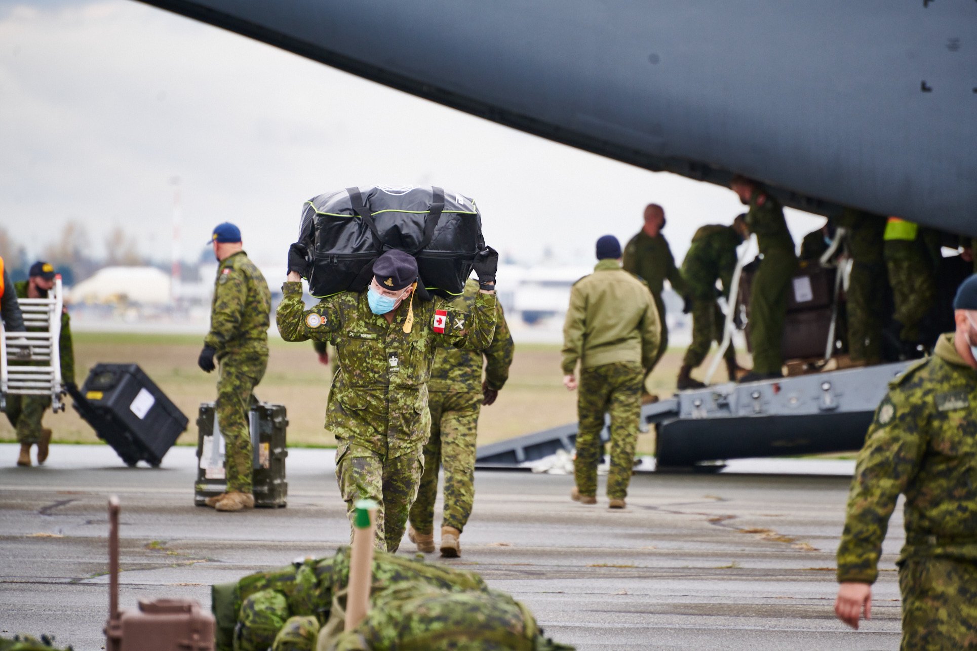 Military personnel in camouflage uniforms working on an aircraft at an airfield, some carrying equipment and wearing masks.