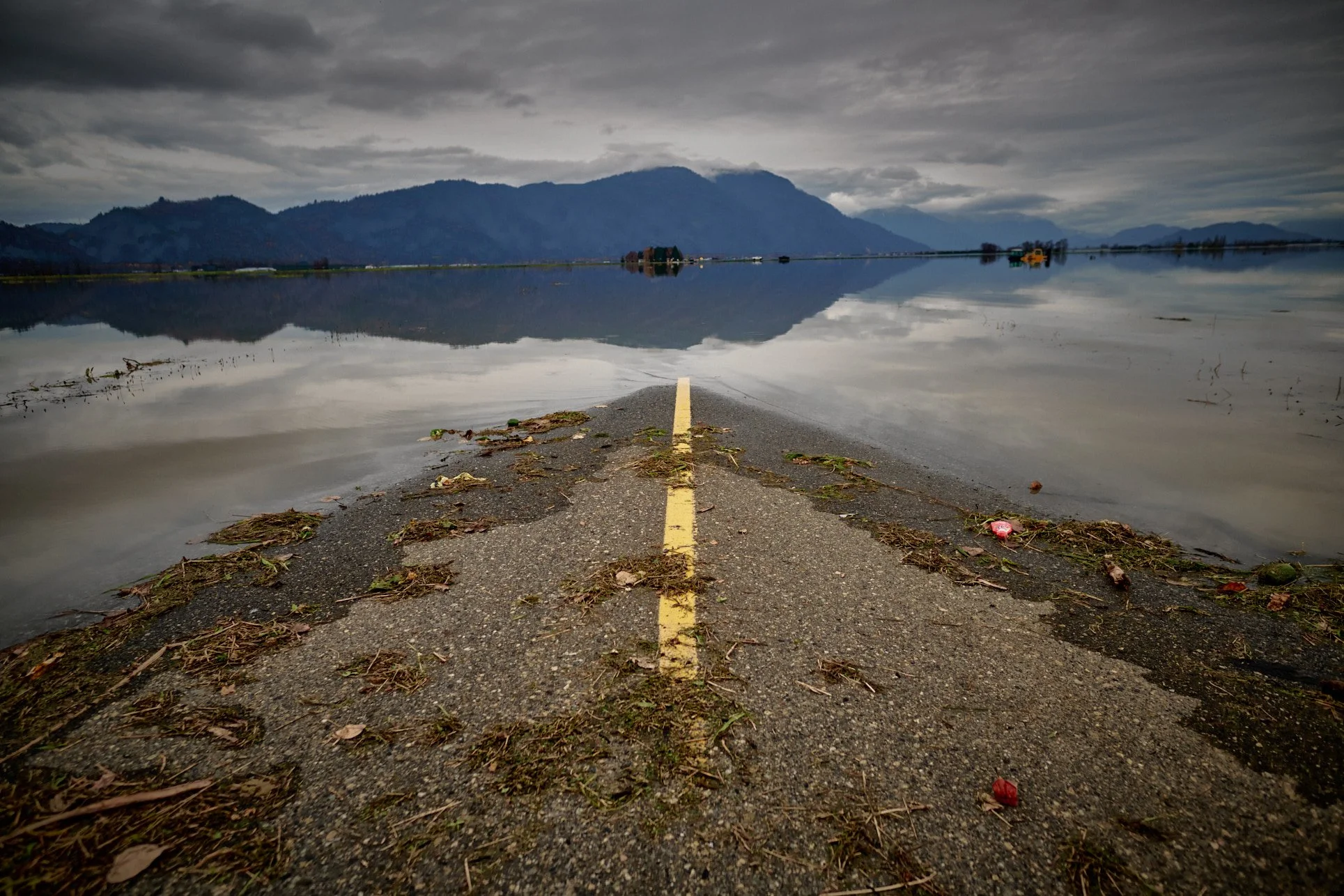 A road extending into a flooded body of water with mountain range in the background and dark cloudy sky overhead.