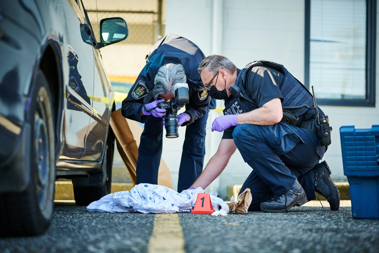 Two police officers investigating a crime scene, examining a body on the ground covered with a white sheet, next to a black car, with evidence markers and photography equipment.