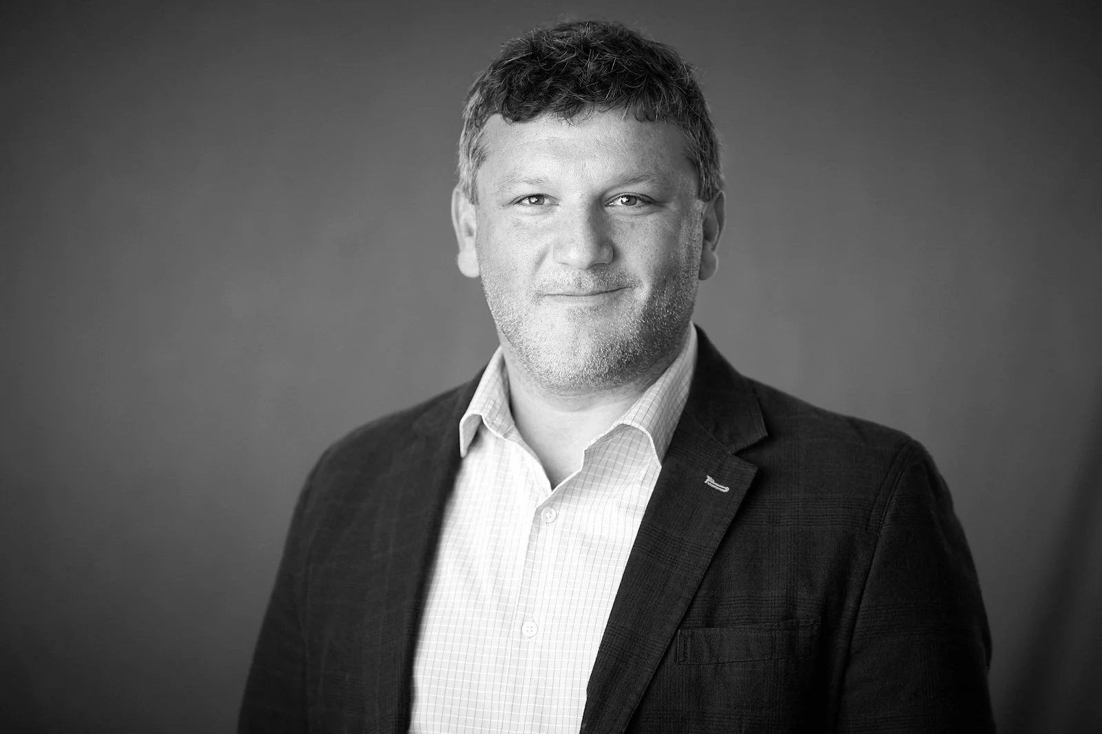 Black and white professional photo of a man with short, curly hair, wearing a blazer over a collared shirt, smiling slightly against a plain background.