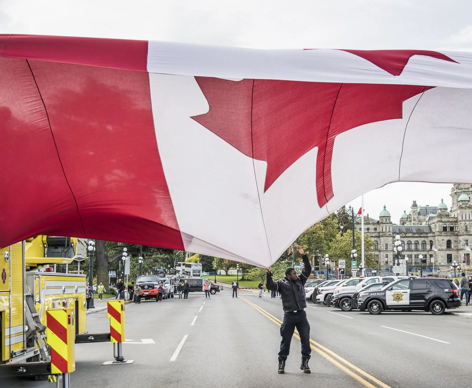 A person holding up a large Canadian flag during a public event, with police cars and historic buildings in the background.